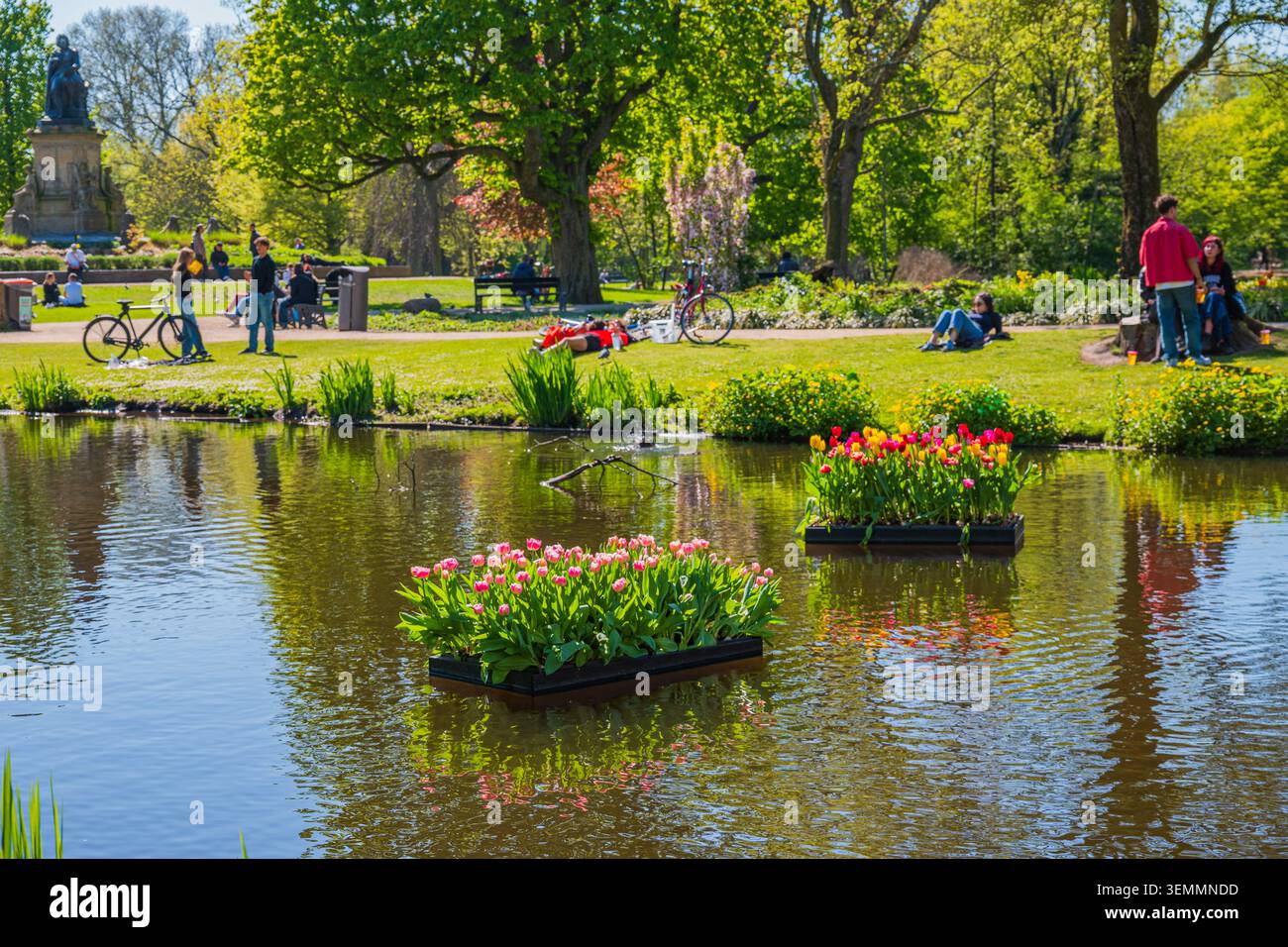 Amsterdam, Paesi Bassi, 22.04.2026 - Vista dello stagno primaverile del Vondelpark di Amsterdam con aiuole galleggianti di tulipani e persone che si rilassano sui prati verdi sotto di su Foto Stock