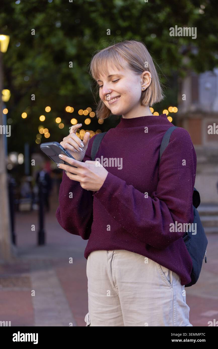 Donna in piedi per strada al crepuscolo che indossa un maglione marrone pantaloni abbronzanti con zaino per smartphone Foto Stock