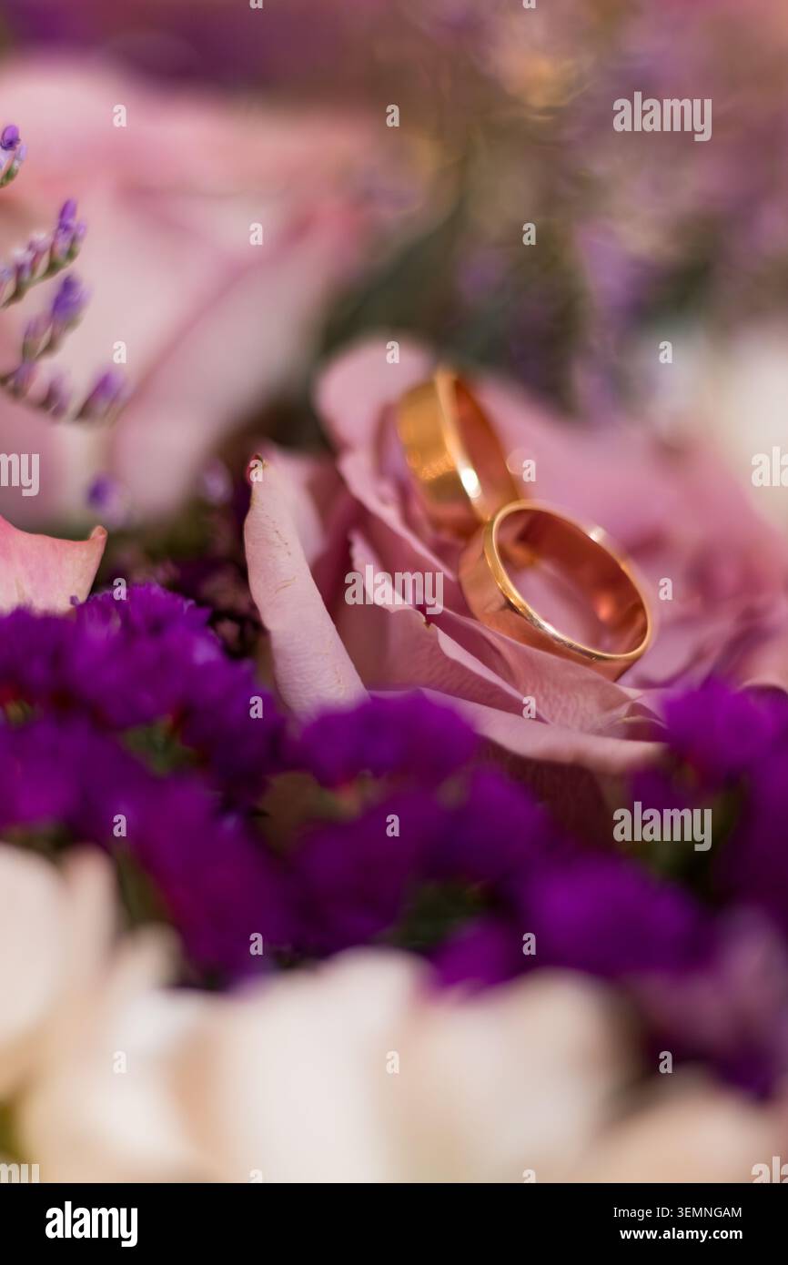 Due fedi nuziali dorate riposano su rose rosa tenui circondate da fiori viola in una composizione floreale. Questa scena cattura un momento di amore e impegno Foto Stock