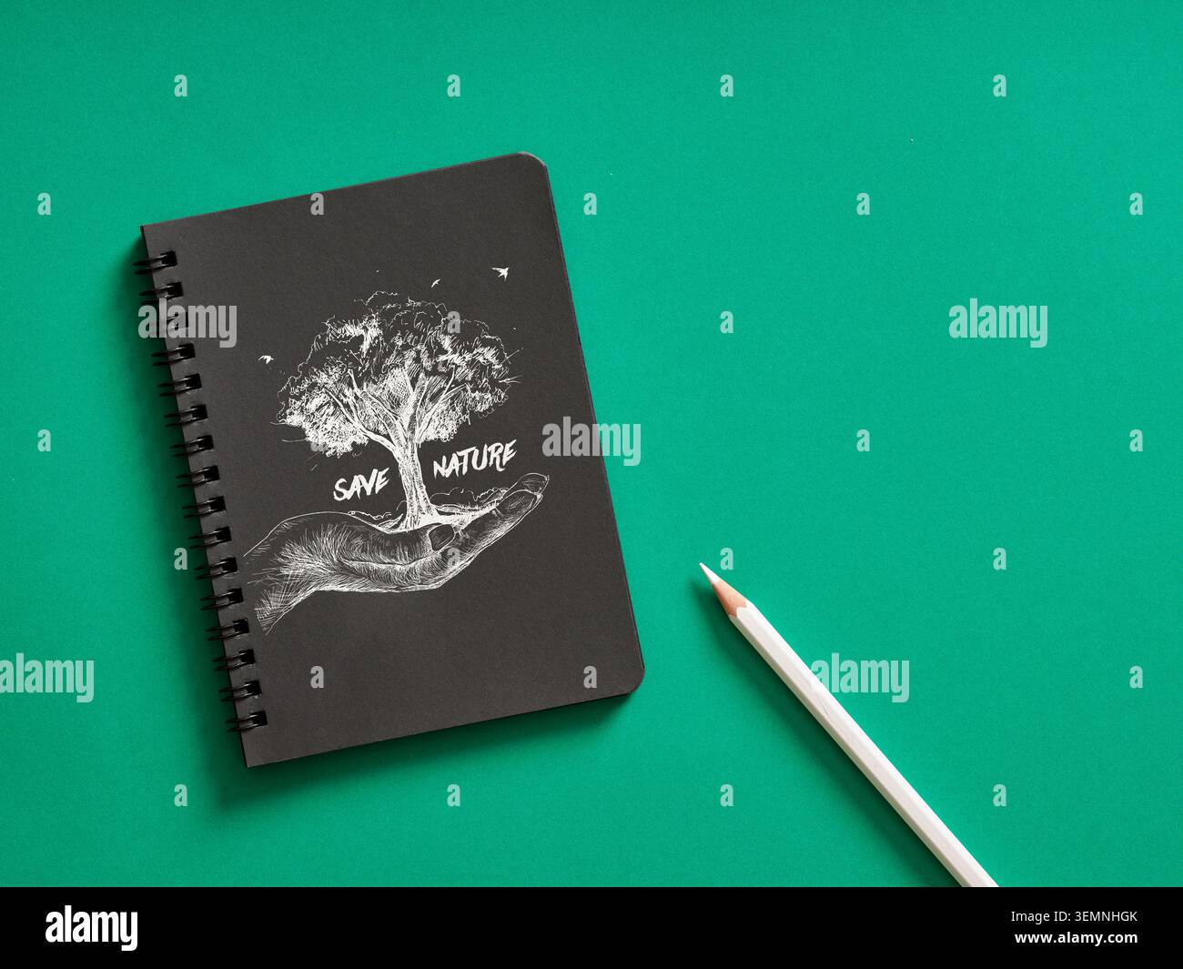 Quaderno a spirale nero con il disegno di un albero in mano e SALVARE il testo NATURALE, posizionato accanto a una matita bianca su sfondo verde Foto Stock