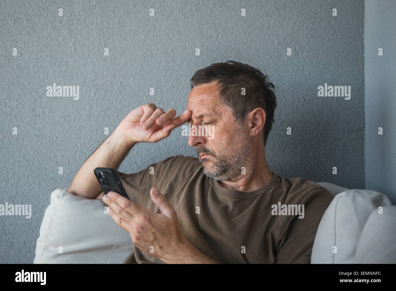 Un uomo di mezza età studia il suo smartphone con un'espressione seria e preoccupata, magari leggendo messaggi o notizie. Abbigliamento casual e sfondo neutro Foto Stock