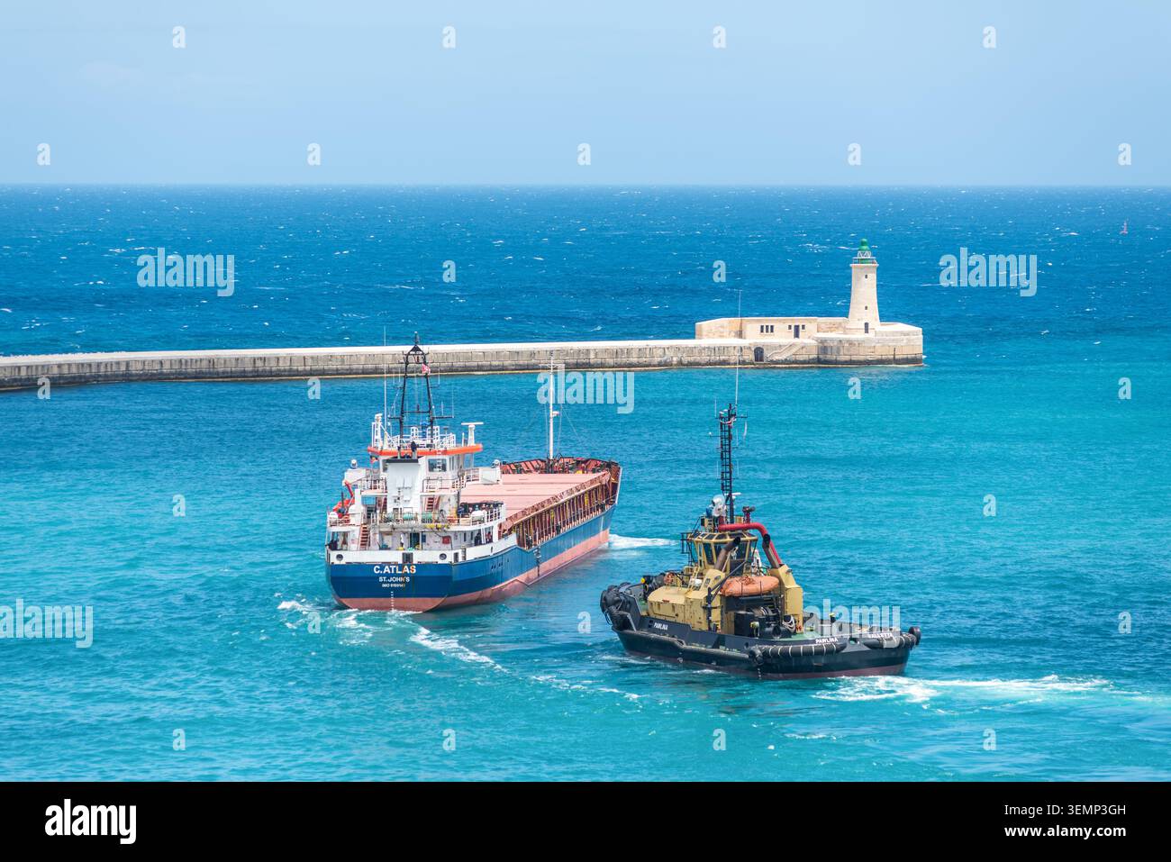 La Valletta, Malta - 16 maggio 2025: Nave da carico e rimorchiatore in barca a vela vicino allo storico ingresso del porto e al faro di la Valletta, Malta, circondato da bricco Foto Stock