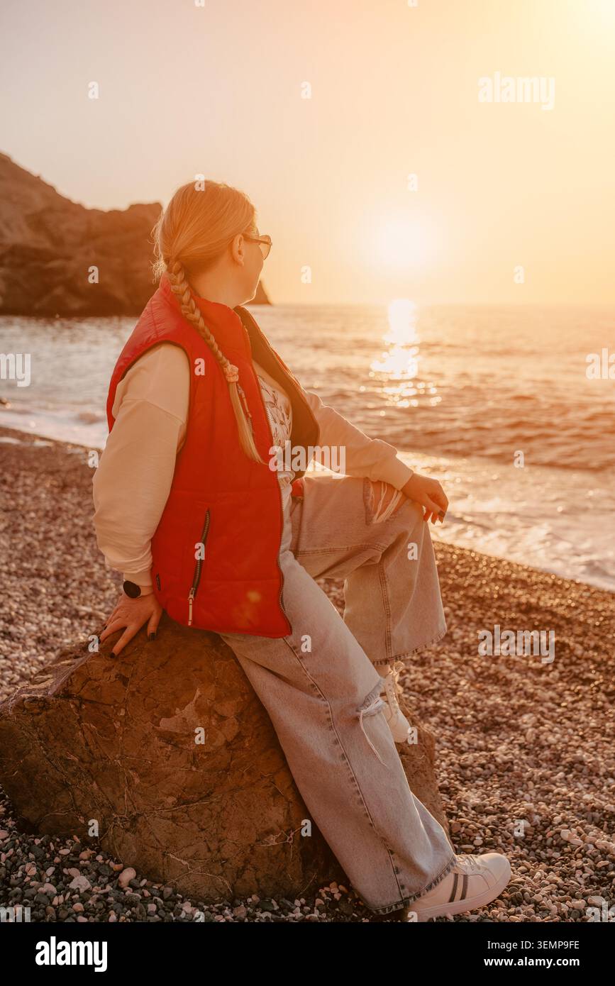 Donna al tramonto sulla spiaggia seduta su una roccia in una spiaggia di ciottoli, osservando il sole dorato che si tuffa sotto l'orizzonte dell'oceano Foto Stock