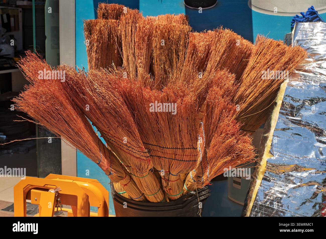 Un pacchetto di tradizionali spazzatrici di sorgo di paglia fatte a mano legate con fili colorati si erge in un secchio nero in un mercato di strada locale. Foto Stock