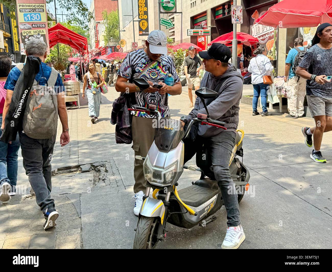Gli uomini guardano un telefono cellulare accanto a uno scooter in Calle Dolores a Chinatown, il centro storico di città del Messico, con pedoni e venditori ambulanti intorno - Immagine stock catturata con smartphone
