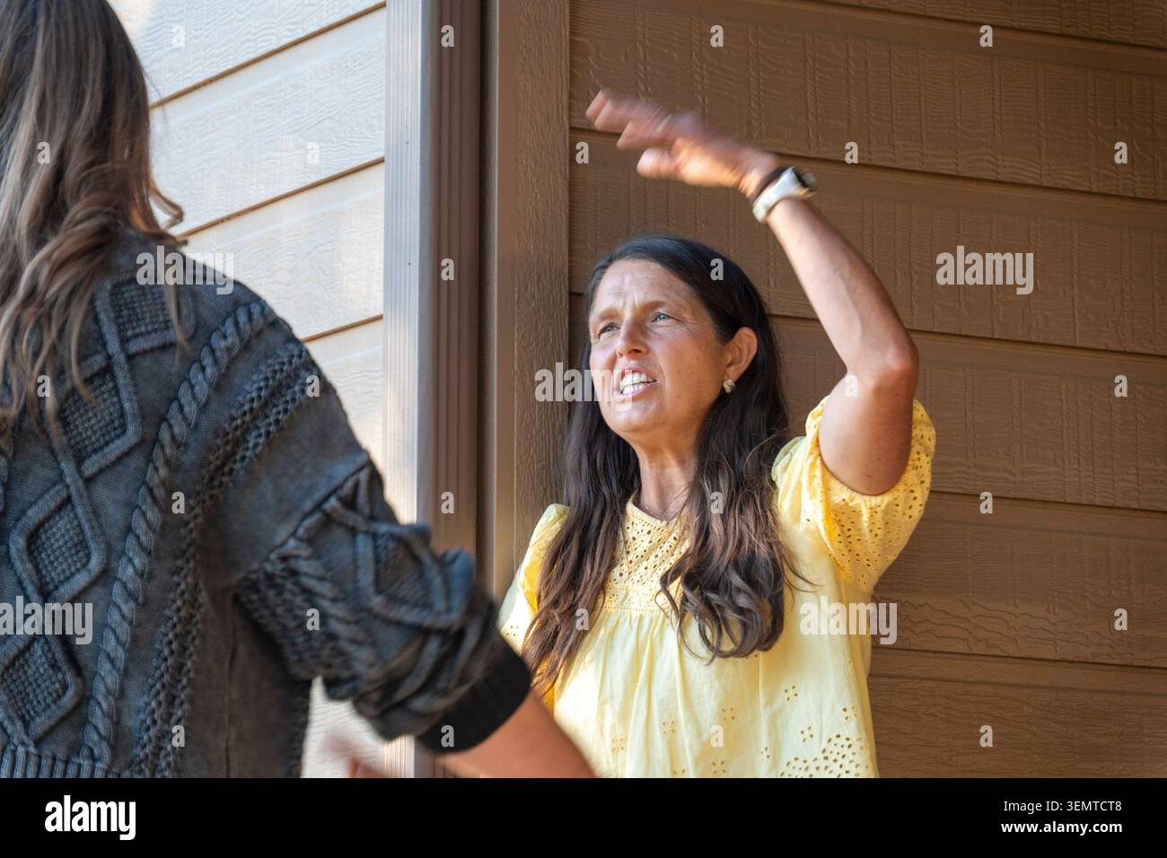 Una scena all'aperto candida che mostra due donne all'ingresso di una casa di legno. Una donna con una camicetta gialla solleva il braccio con un movimento gestuale, mentre la Foto Stock