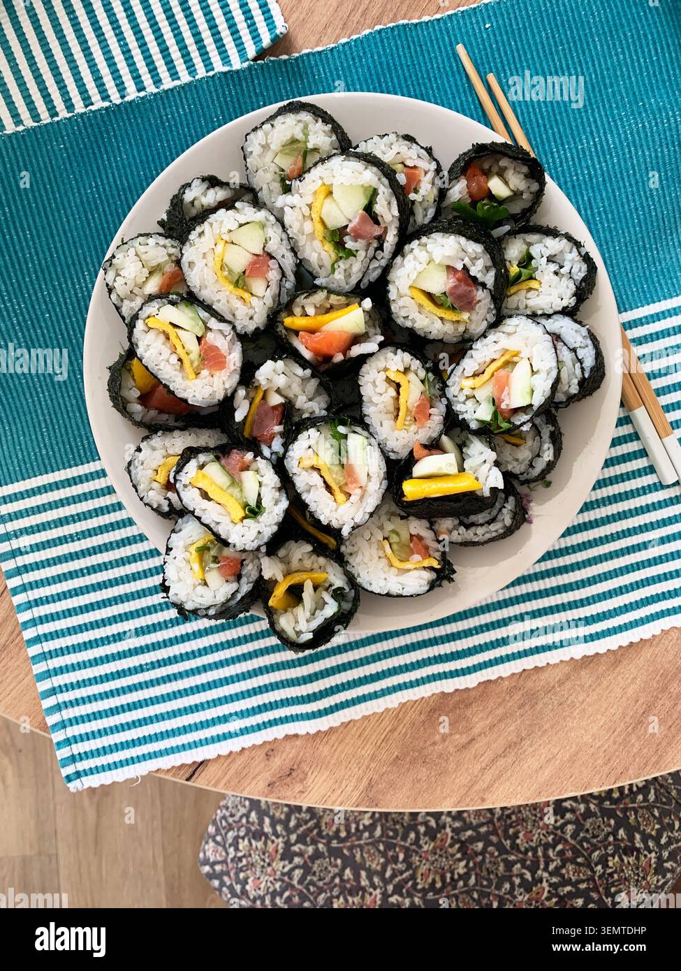 Gli involtini di sushi sono disposti ordinatamente su un piatto bianco. I panini contengono cetriolo, pesce rosso e uova. Sono serviti con bacchette su un tappeto blu e verde Foto Stock