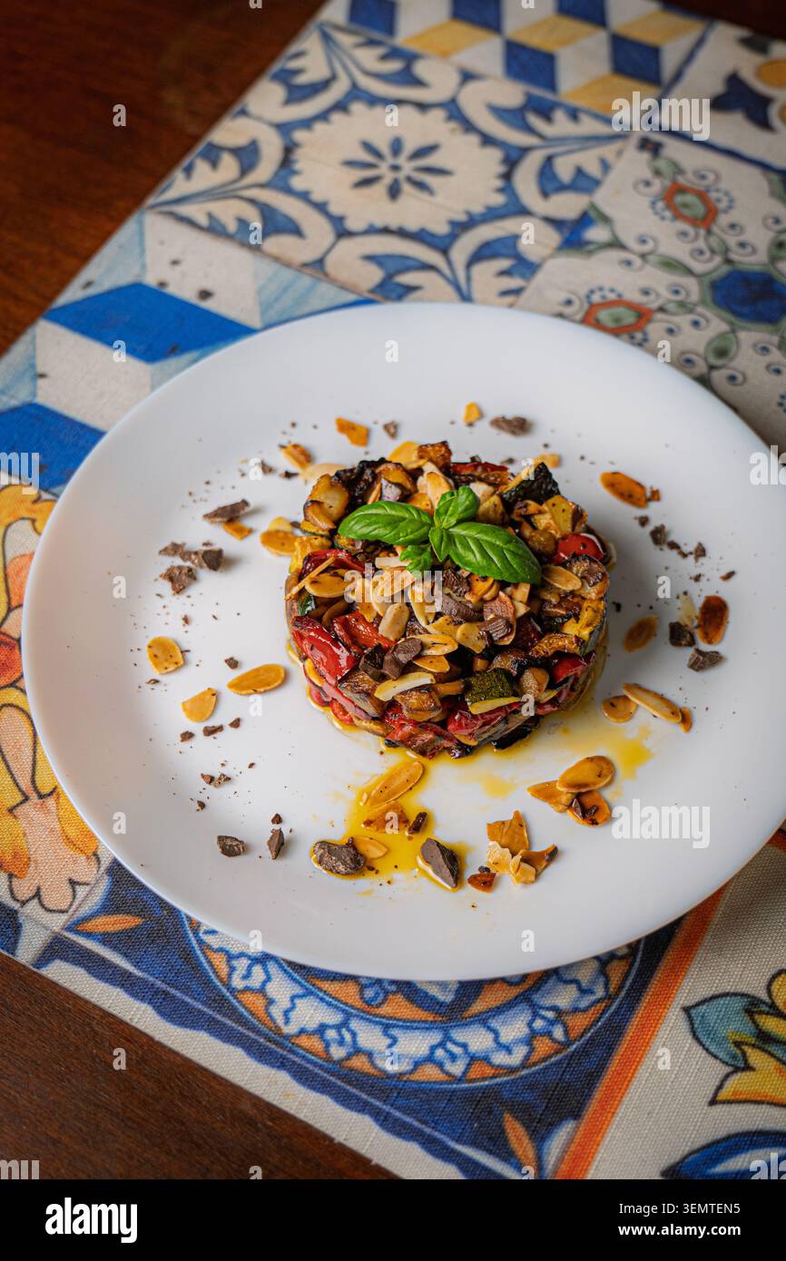 Una caponata siciliana elegantemente placcata, presentata come una pila rotonda stampata di verdure arrostite, tra cui melanzane, zucchine e pepe rosso, topp Foto Stock