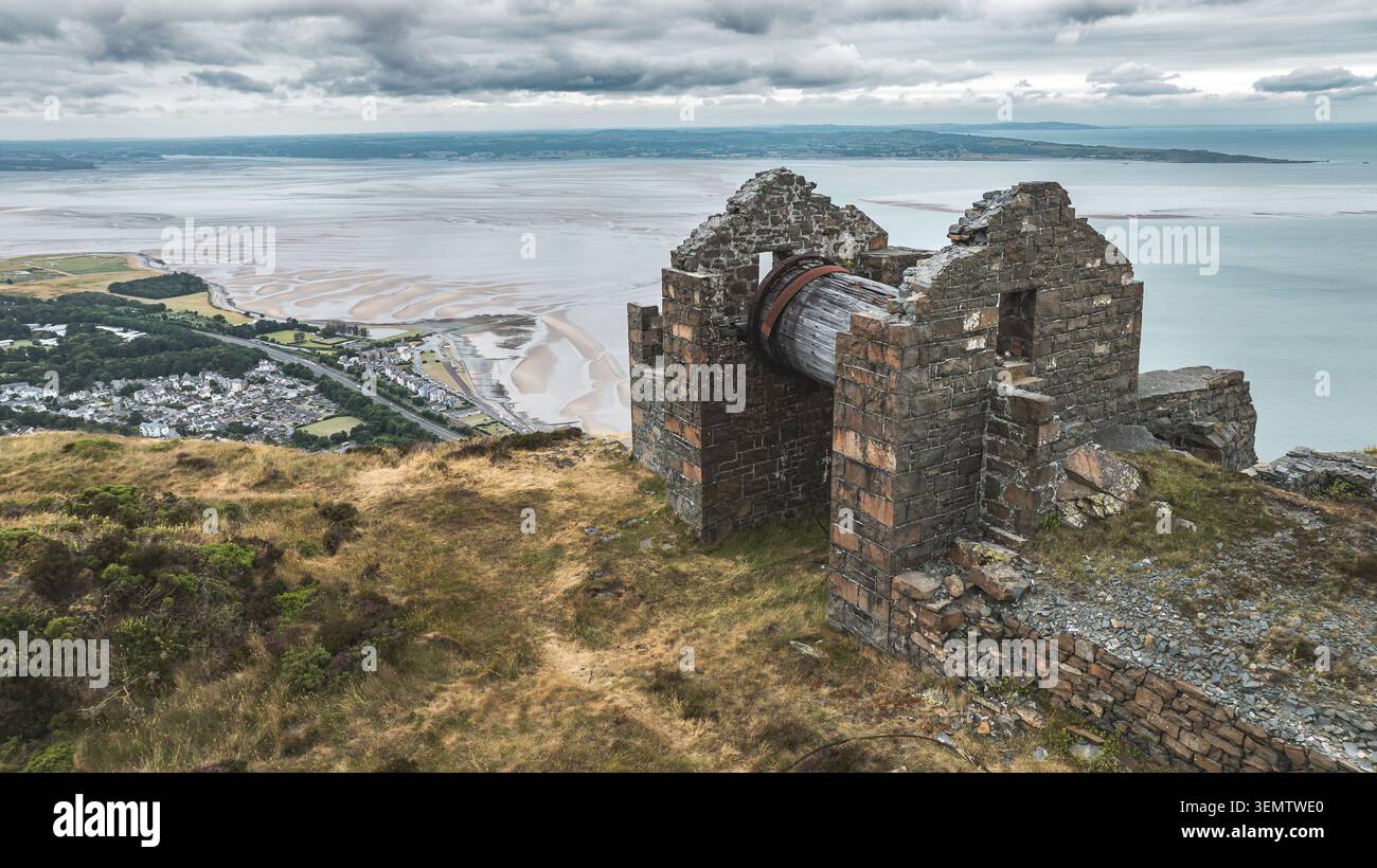 Un edificio abbandonato in pietra con una grande ruota di legno sorge su una collina erbosa sopra una cittadina di mare e un ampio estuario sabbioso sotto un cielo nuvoloso Foto Stock