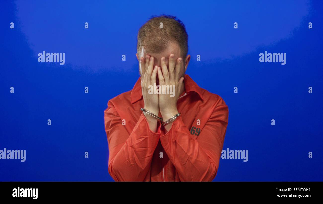 Tuta da giovane in prigione arancione con mani che coprono il viso e manette in metallo in studio contro il muro blu; rimorso. Foto Stock