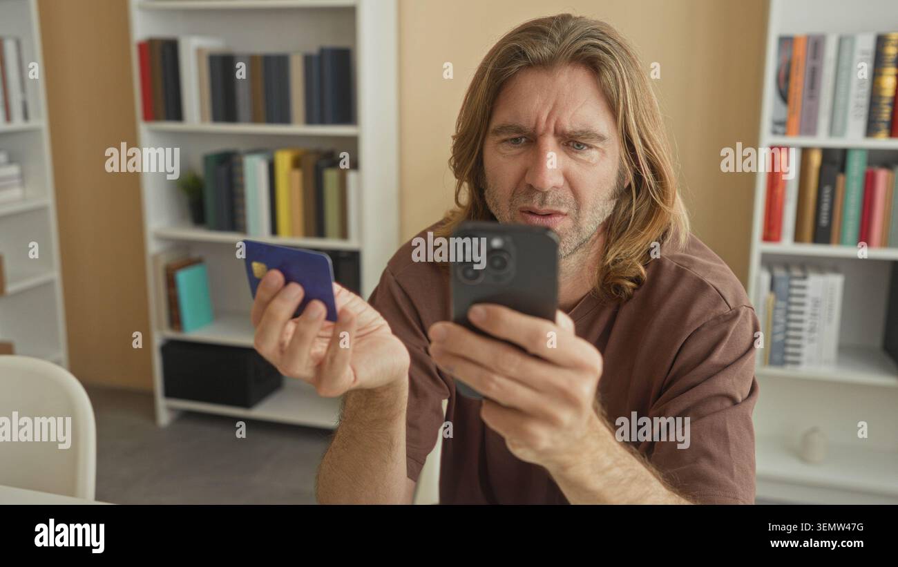 Uomo con smartphone e carta di credito, strizza sullo schermo mentre controlla i dettagli della carta in un edificio di studio; frustrazione. Foto Stock