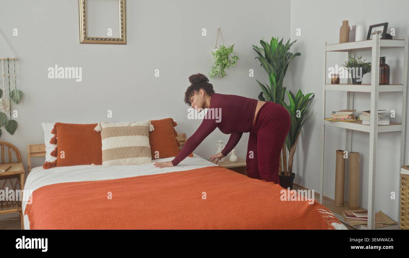 Donna che nasconde una coperta arancione sul letto con le mani e i cuscini liscianti mentre si piega vicino allo scaffale in una camera da letto dell'edificio; calma routine. Foto Stock