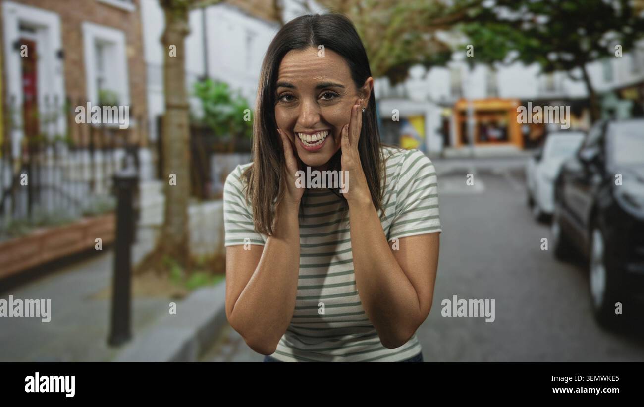 Donna che tocca le guance con le mani, sorride e guarda piacevolmente sorpreso in strada con auto parcheggiate e vetrine; gioia di festa. Foto Stock