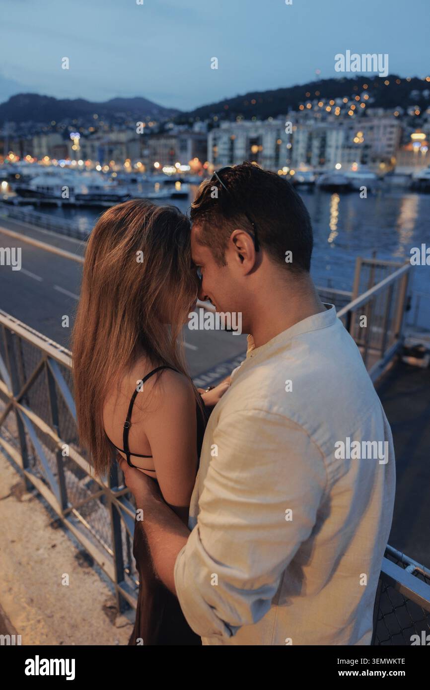 Le luci soffuse al crepuscolo illuminano il paesaggio urbano lontano. La coppia sta vicino, le facce quasi toccanti, intime e tranquille. Si guardano negli occhi dell'altro, sh Foto Stock
