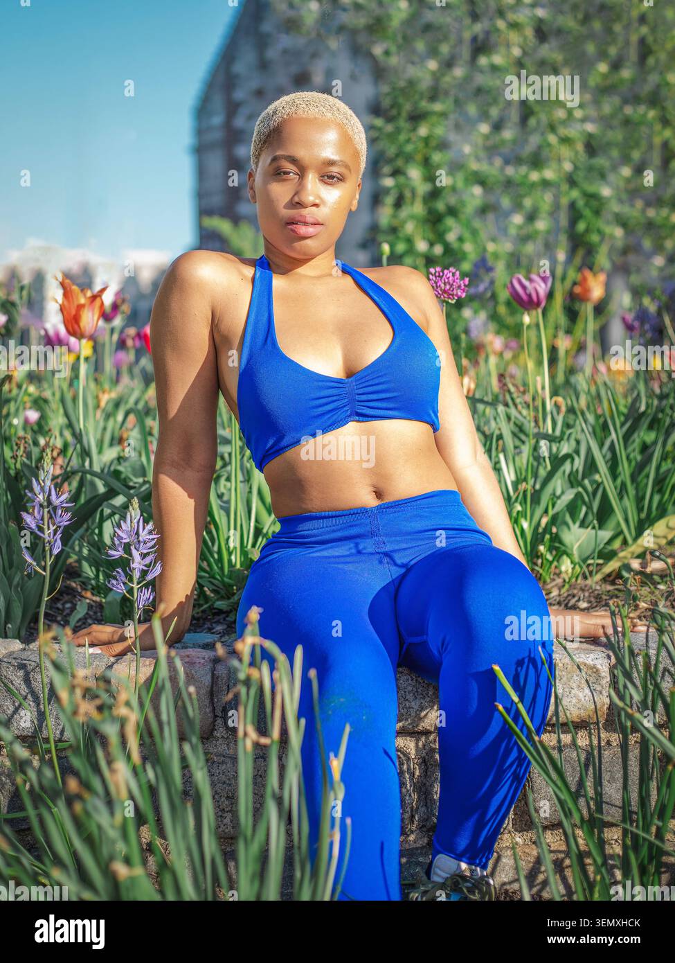 Attraente giovane donna nera con capelli biondi corti in posa con un vivace top sportivo blu e leggings a vita alta abbinati a un flo lussureggiante Foto Stock