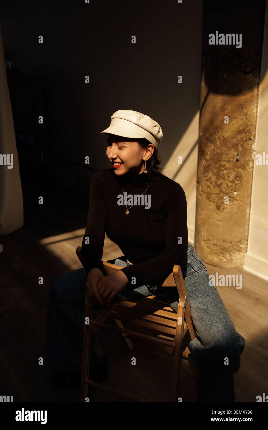 Una donna è seduta su una sedia di legno. Indossa un cappello e si siede in un'area illuminata dal sole di una stanza. La scena mostra una luce soffusa e uno sfondo semplice con un co Foto Stock