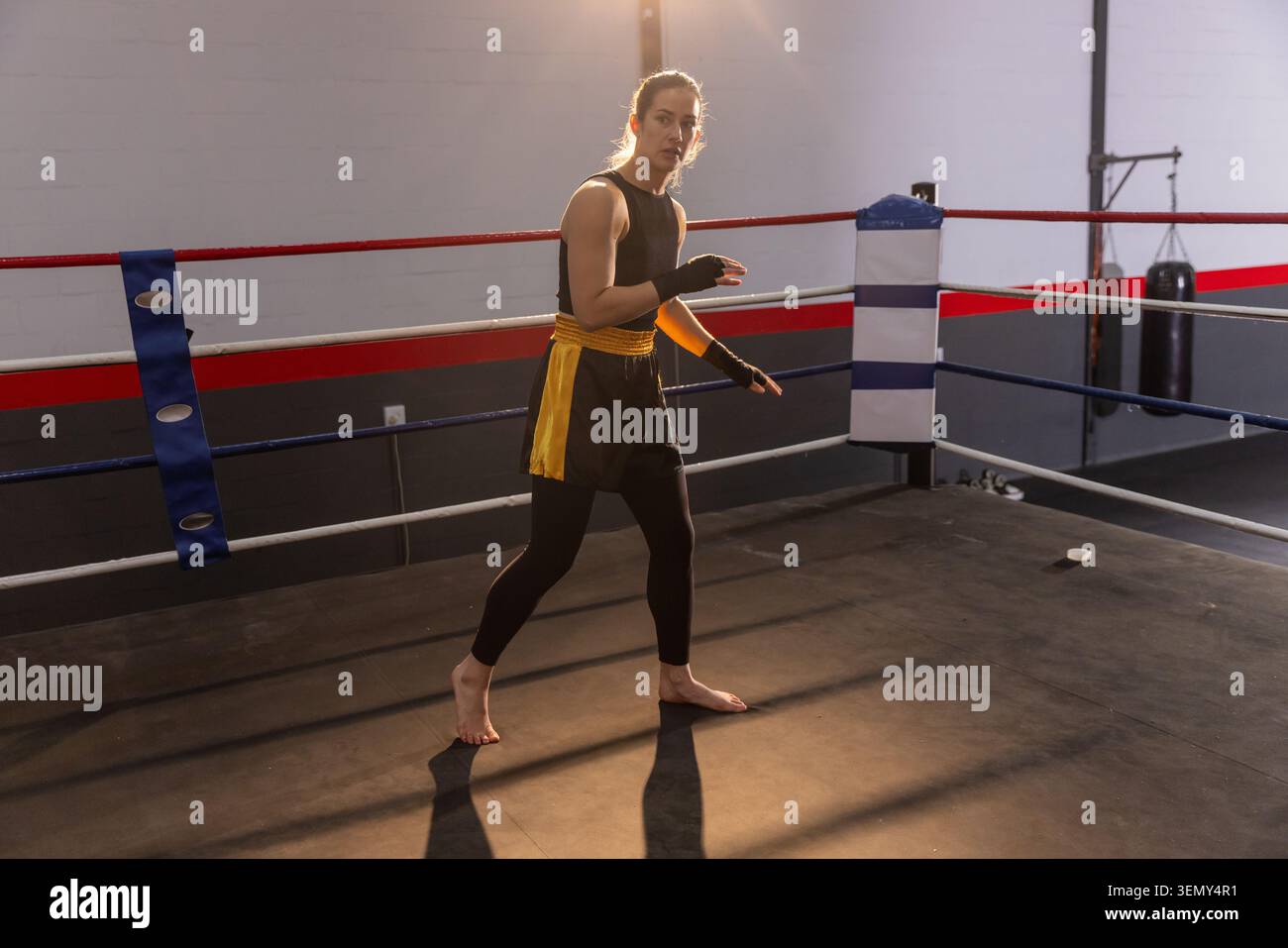 Pugile donna in piedi sul ring in palestra, indossa pantaloncini neri gialli, tenendo le mani avvolte Foto Stock