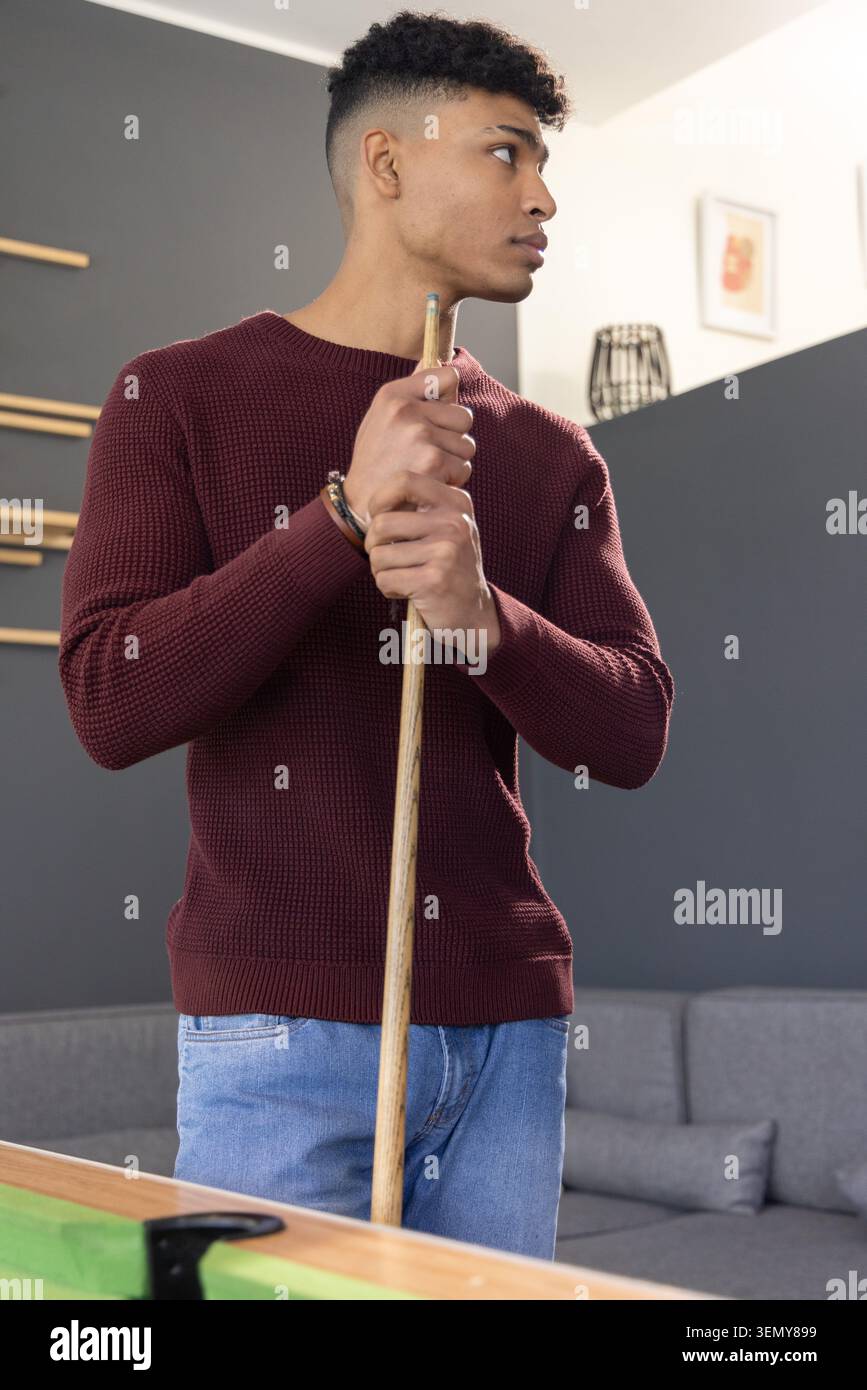 Uomo afro-americano in piedi al tavolo da biliardo a casa tenendo spunto indossando maglione marrone e braccialetti Foto Stock