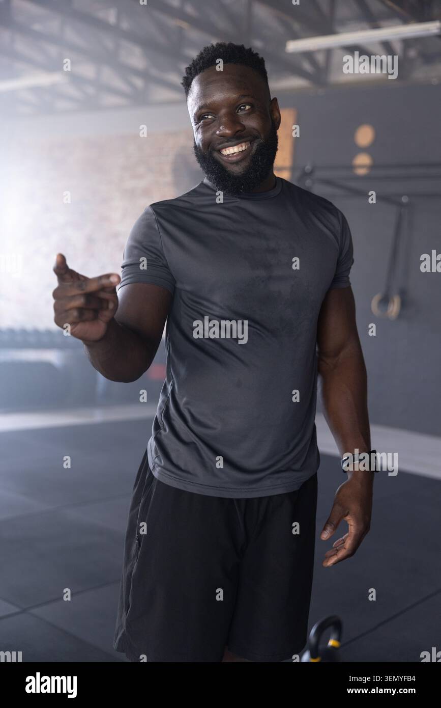 Uomo afro-americano in palestra sorridente, punta, indossa camicia aderente, campanello e anelli Foto Stock
