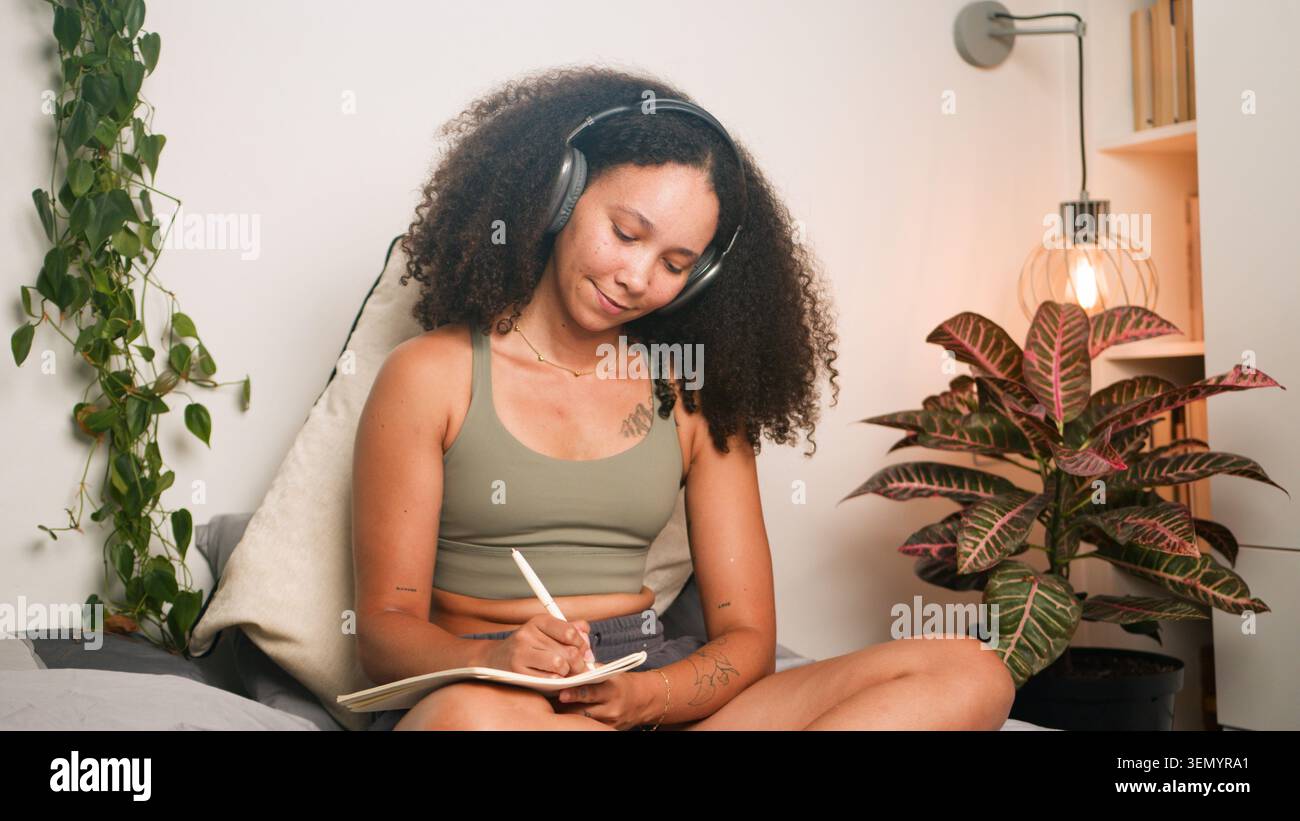 Donna nera che scrive a casa indossa le cuffie nell'accogliente camera da letto con piante e lampada Foto Stock
