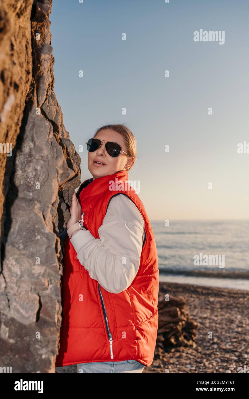 Gilet rosso spiaggia donna che indossa occhiali da sole e si appoggia su una roccia costiera durante un'ora d'oro al tramonto con sfondo marino Foto Stock