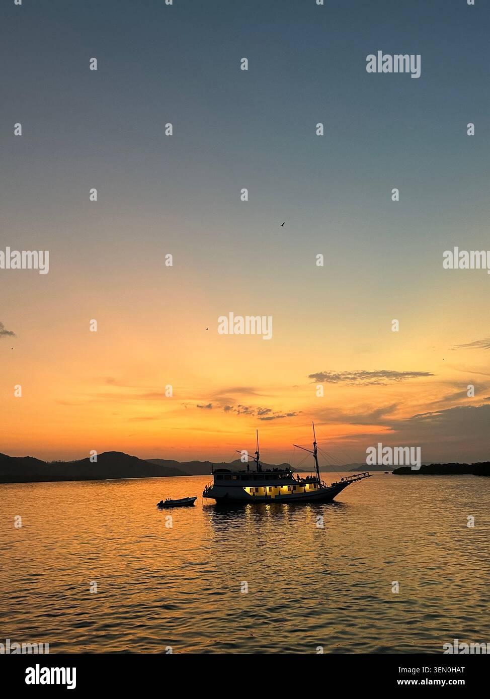 Atmosphera pirata: una barca tradizionale phinisi ancorata nelle acque dorate del tramonto, Parco Nazionale di Komodo, Indonesia - Immagine stock catturata con smartphone