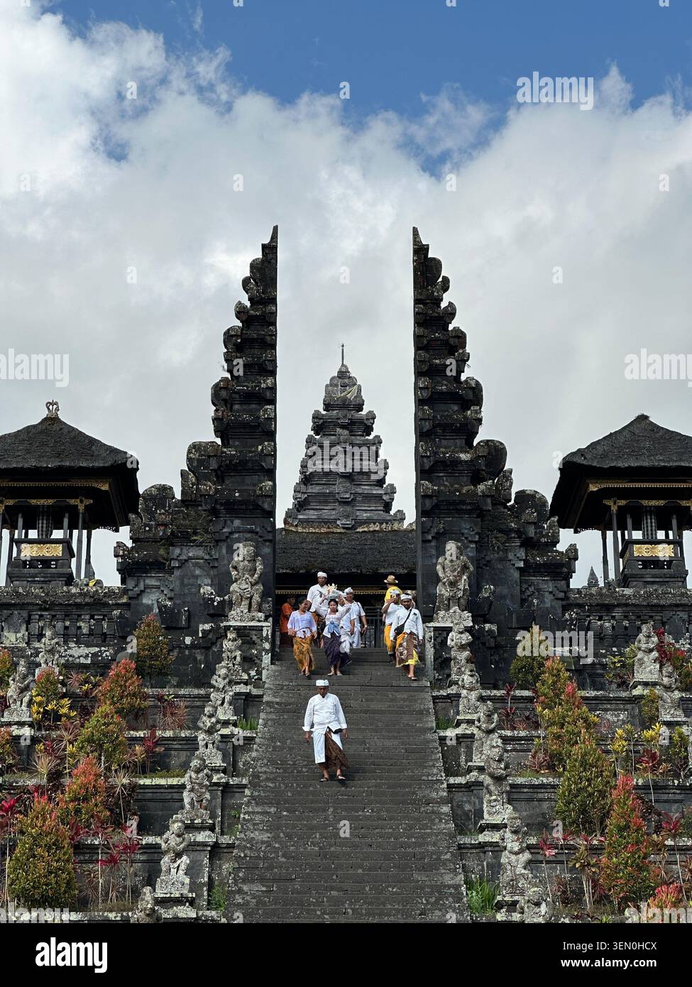 I visitatori salgono una maestosa scalinata in pietra tra porte divise che si innalzano in un tempio balinese fiancheggiato da fiori - Immagine stock catturata con smartphone
