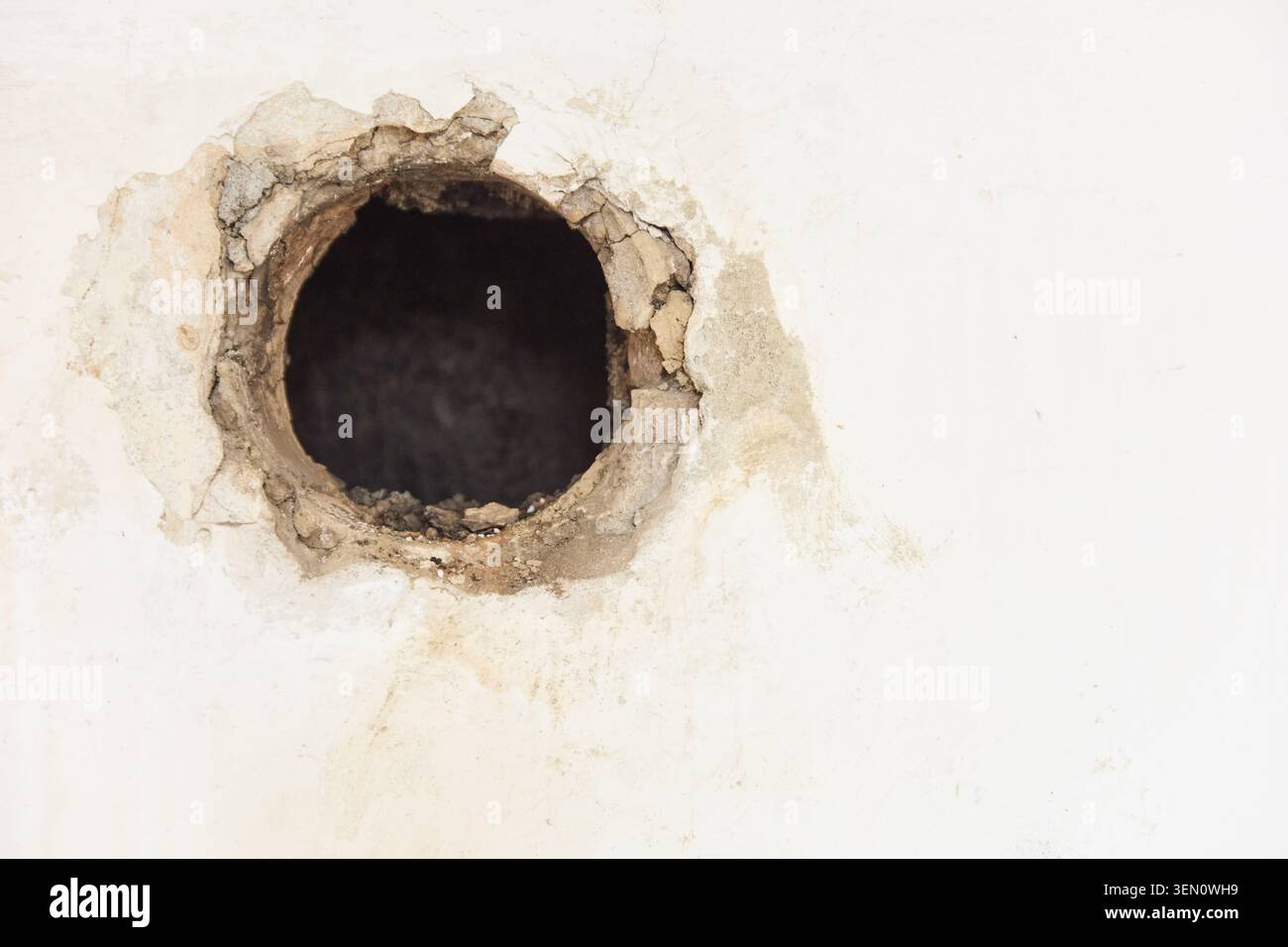 Vista ravvicinata di un foro circolare ruvido in una parete in gesso danneggiata, che indica un progetto di ristrutturazione o demolizione in un vecchio condominio. Foto Stock