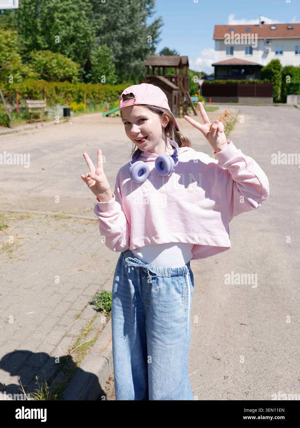 Ragazza alfa gen di tendenza con cappuccio rosa Crop Top che mostra un segno di pace. Indossa cuffie wireless viola in posa sulla strada soleggiata. Via della giovinezza Foto Stock