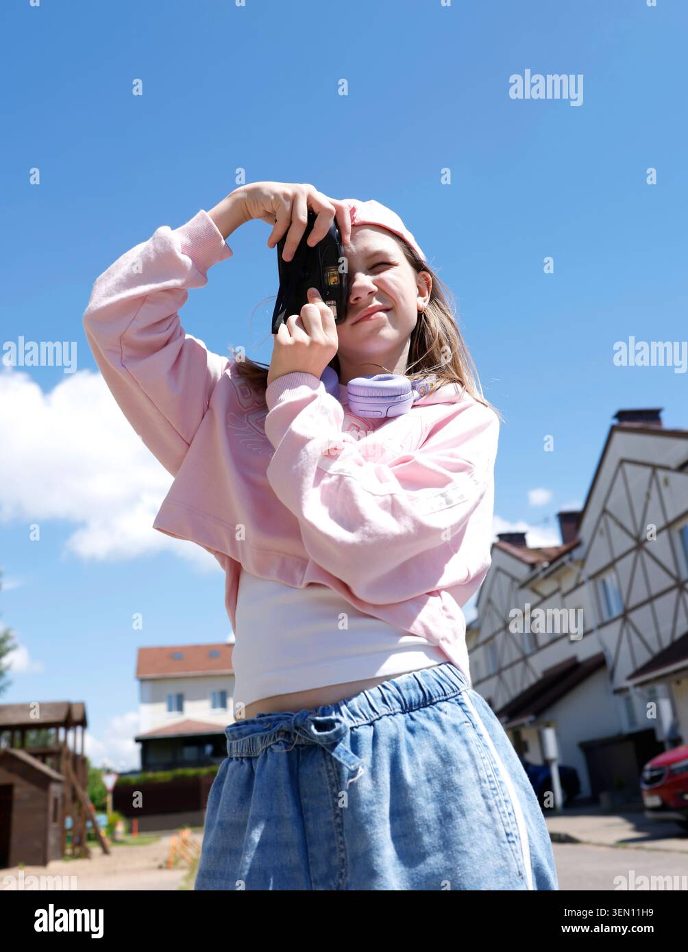 Ragazza alfa di generazione alla moda che scatta foto con una fotocamera vintage all'aperto. Una felpa con cappuccio rosa che regge la pellicola retrò contro il cielo blu. Hobby giovanile e.. Foto Stock