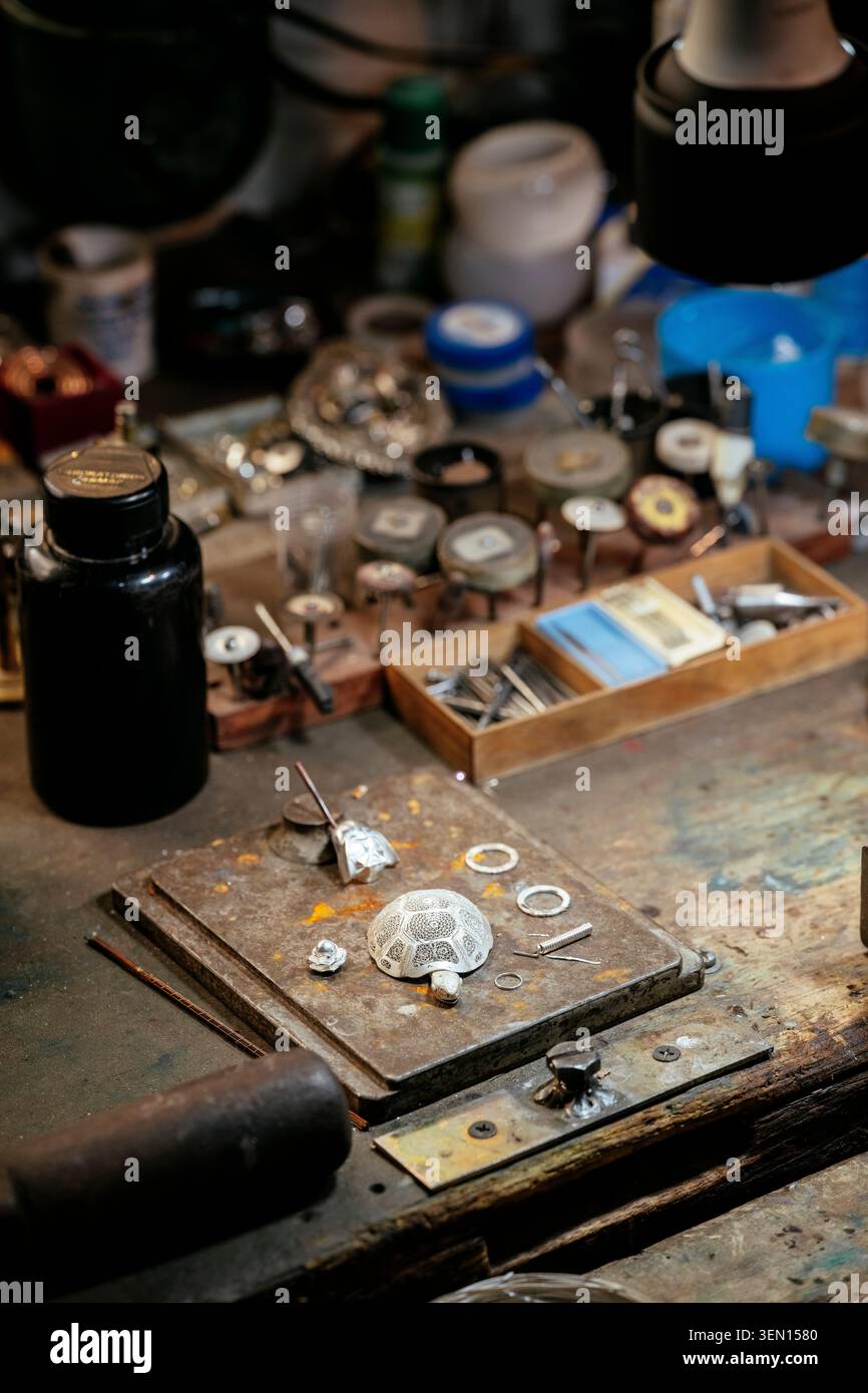 Un banco da lavoro per gioiellieri ingombrante con utensili, pezzi di metallo e una lampada luminosa sopra la testa. Foto Stock