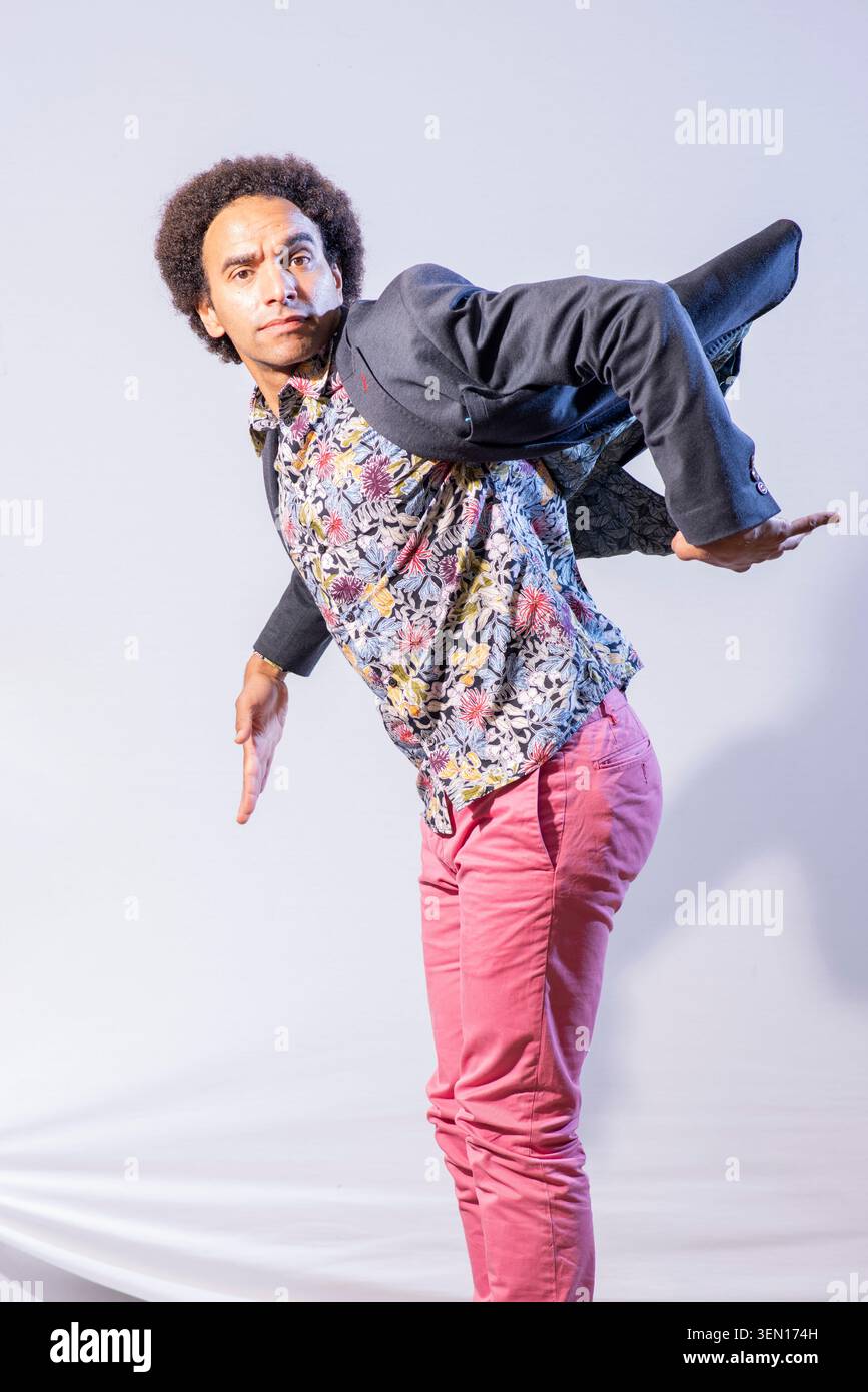 Joseph Coelho si trova in uno studio, indossando una camicia floreale e pantaloni rosa. Posa con il suo blazer aperto e le braccia allungate, mostrando una posizione giocosa Foto Stock