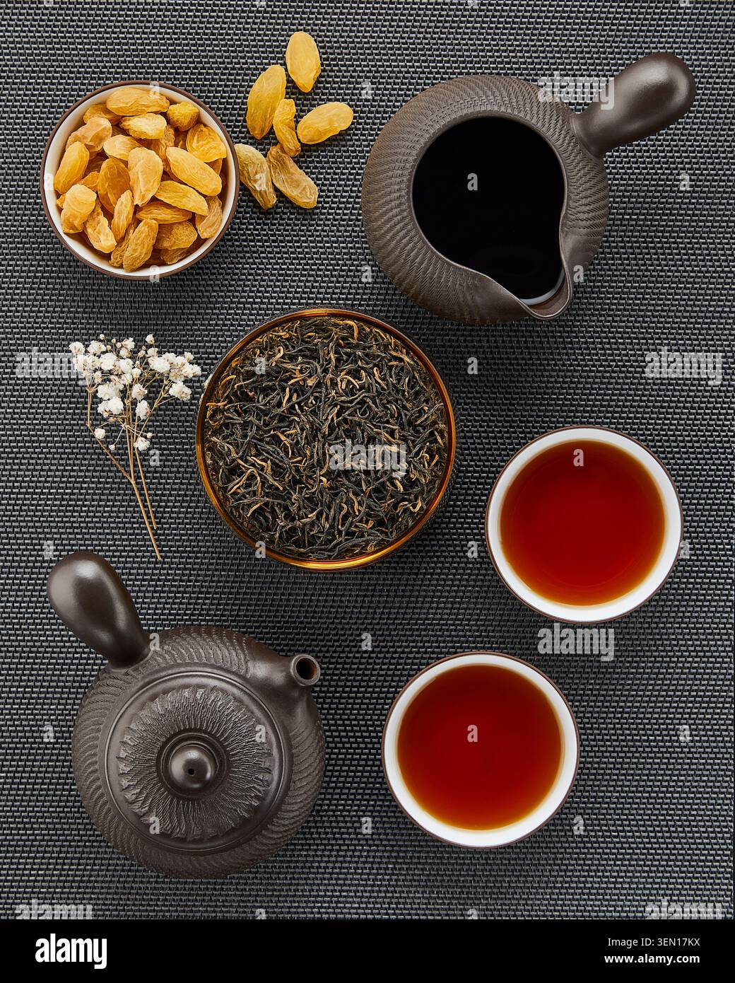 Vista dall'alto del tè nero cinese con tè a foglia sciolta, teiera, caraffa, tazze da tè e uva passa dorata su sfondo tessuto scuro. Foto Stock