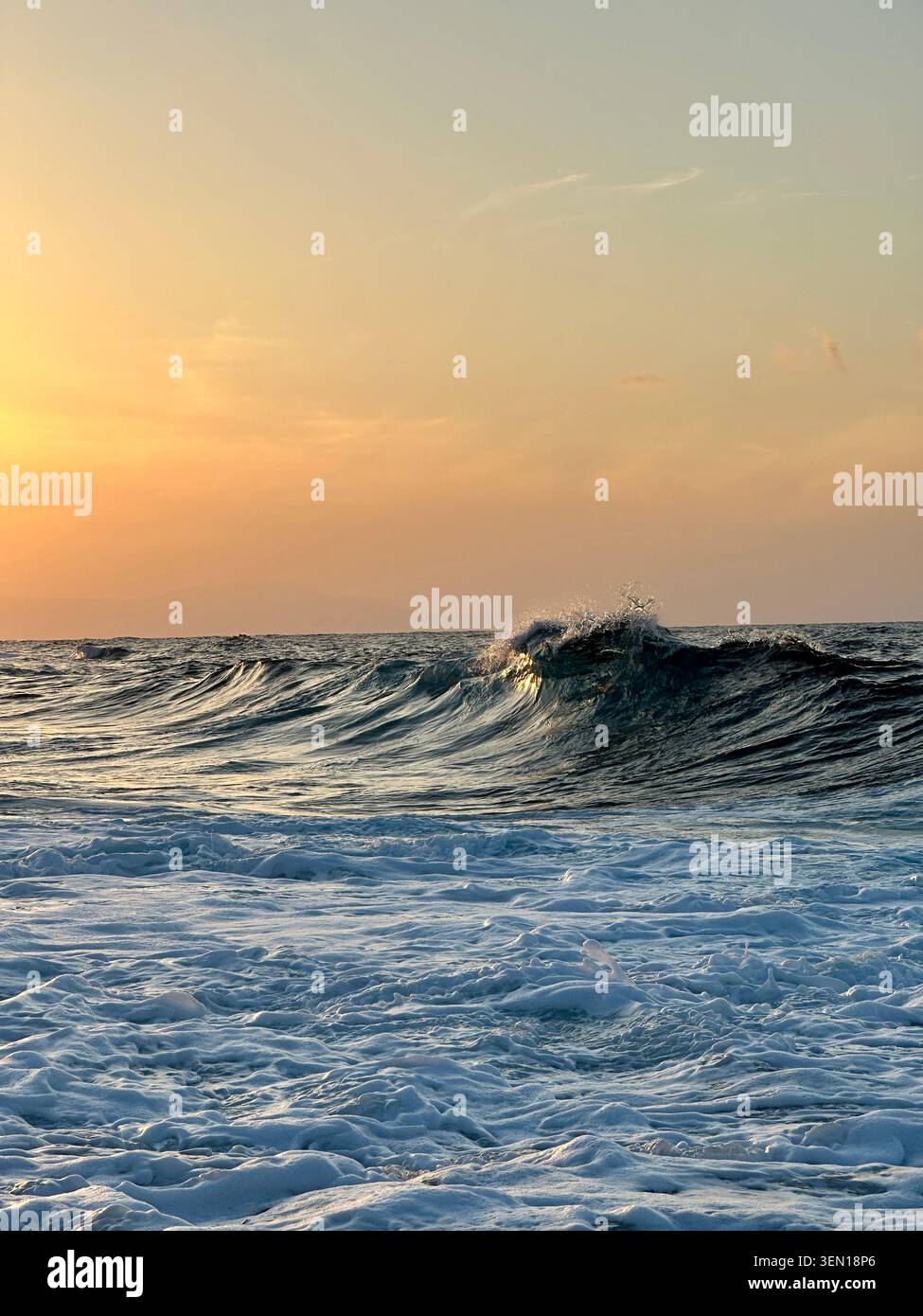 Un'onda rotolante si solleva dal mare blu-argentato sotto un tranquillo cielo pastello al tramonto dipinto con luce arancione. Foto Stock