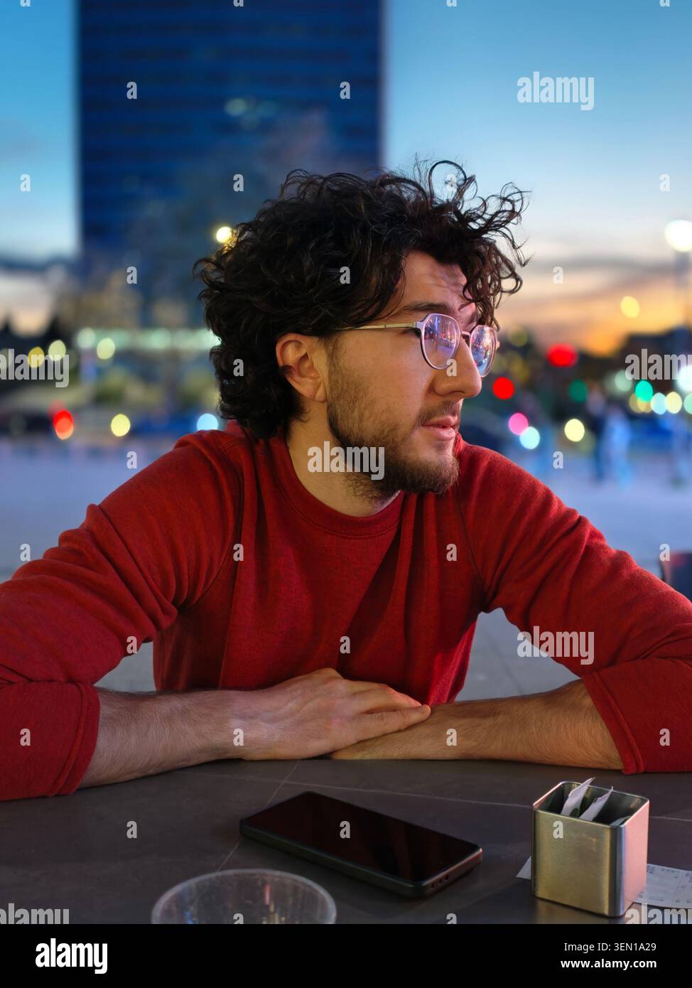 Giovane uomo in maglione rosso con occhiali seduto al tavolo all'aperto nel ritratto serale della città Foto Stock