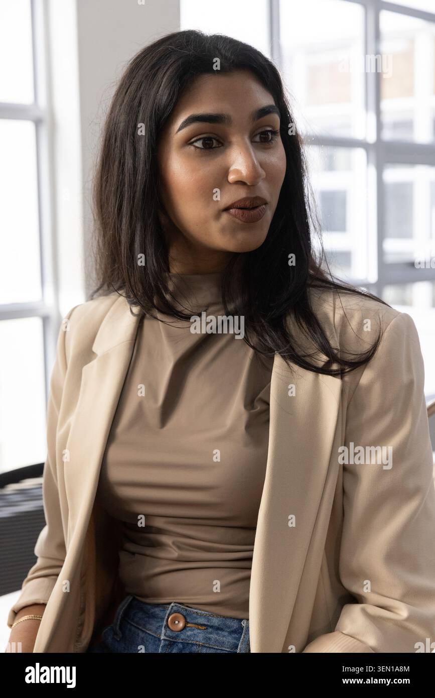 Donna indiana seduta in un ufficio luminoso con blazer beige, top taupe, jeans blu, braccialetto sottile Foto Stock