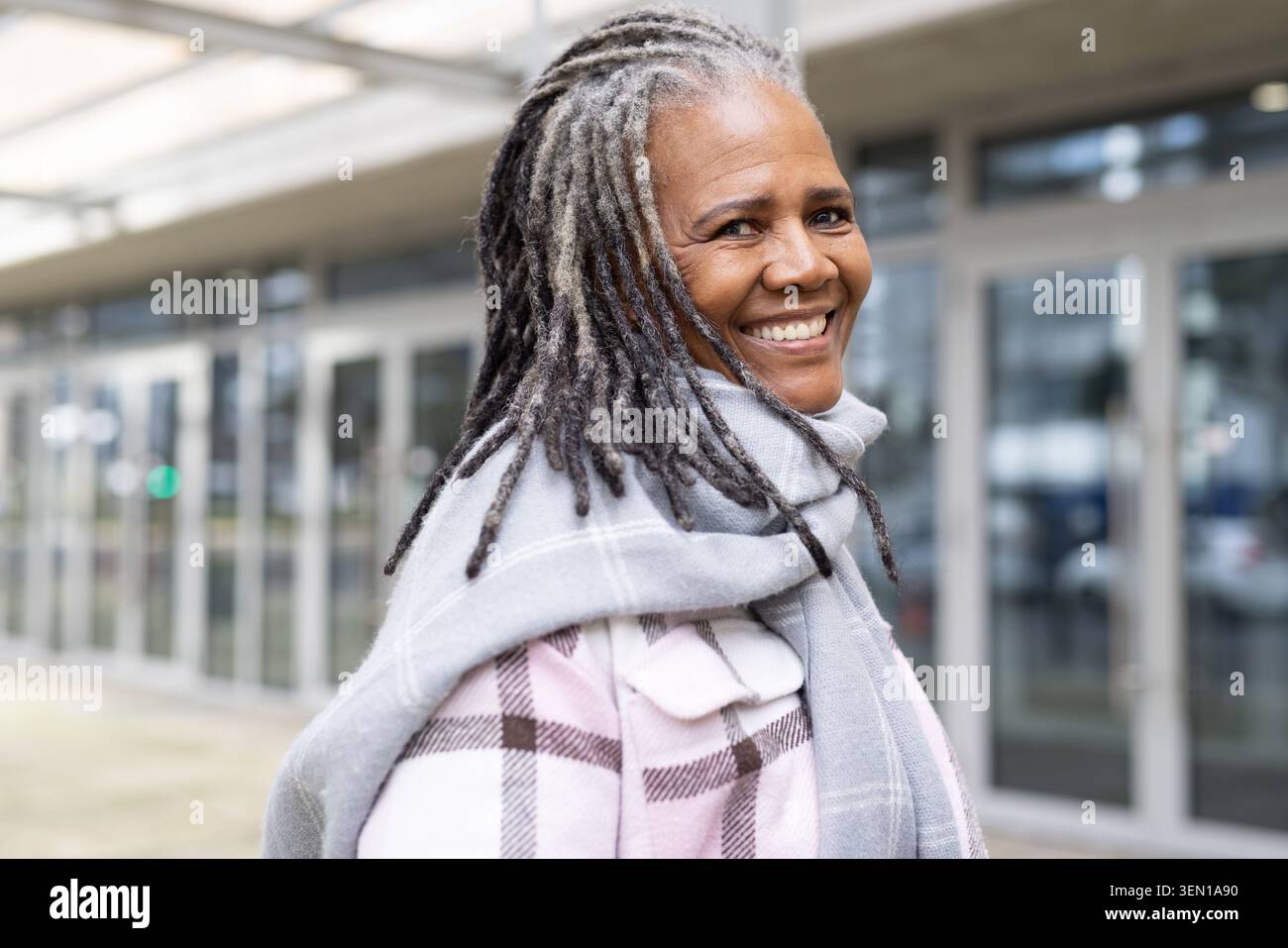 Donna anziana sorridente in piedi vicino all'ingresso moderno con un cappotto a quadri pastello sciarpa grigia lunga teme Foto Stock
