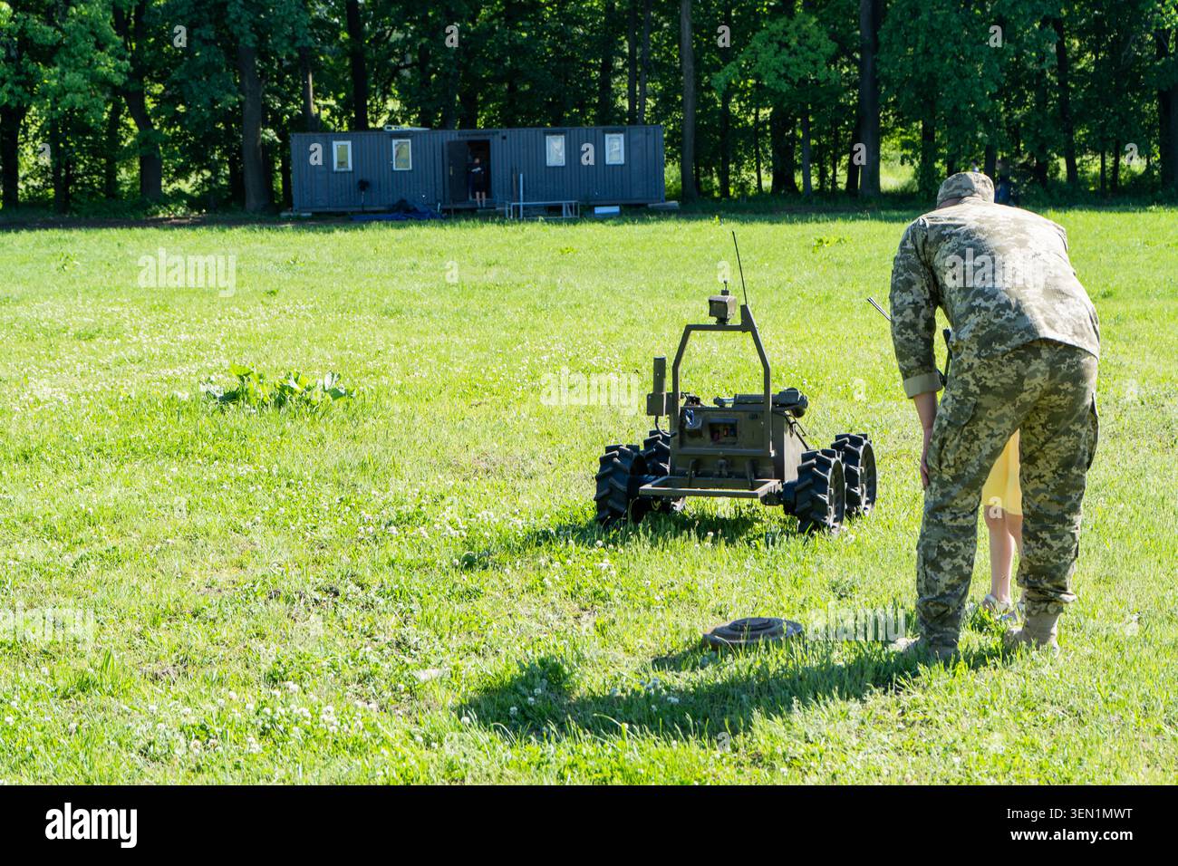 L'ufficiale militare dimostra un veicolo di terra senza equipaggio monitorato a distanza sul Green Grass Field. Sminamento dei terreni nel campo agricolo. Il sapper sta funzionando. Vista posteriore di un uomo Foto Stock