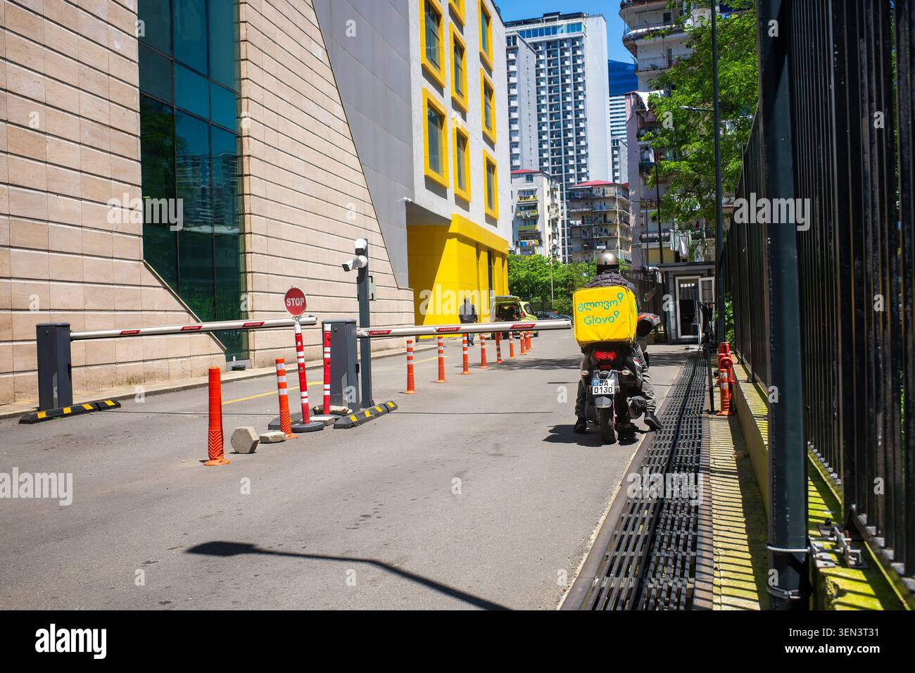 Batumi, Georgia - 3 giugno 2026: Corriere con servizio di consegna di cibo a bordo di uno scooter con una scatola gialla Glovo presso le braccia automatiche della barriera di controllo. Foto Stock