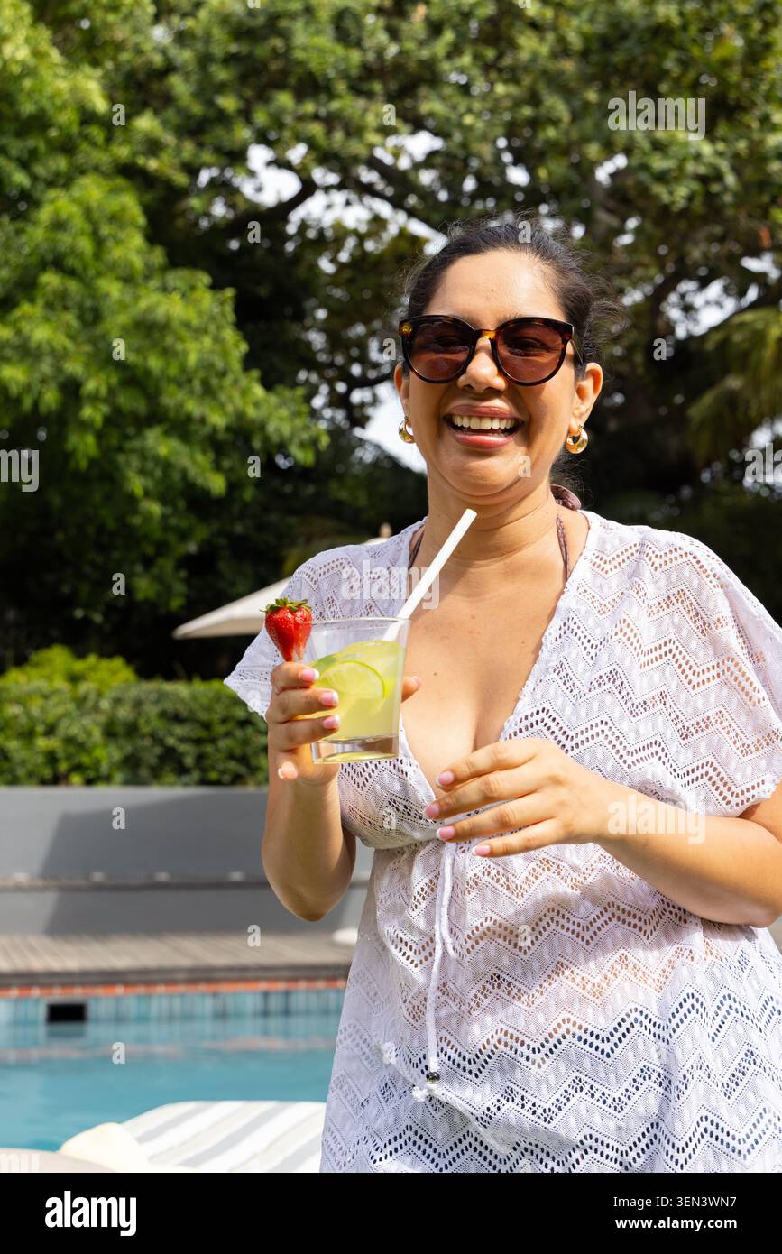 Donna che indossa un copriletto all'uncinetto ridendo mentre tiene la tazza con lime, paglia e fragola a bordo piscina Foto Stock