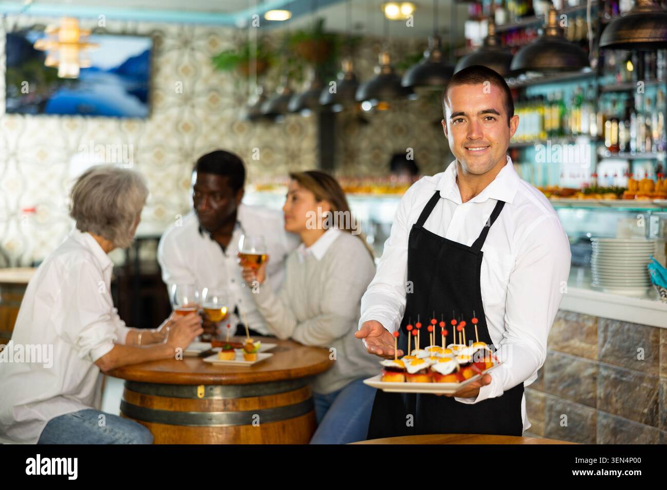 Sorridente cameriere latino nel grembiule che serve antipasti, tapas, spuntini sugli spiedini per persone diverse nel pub Foto Stock