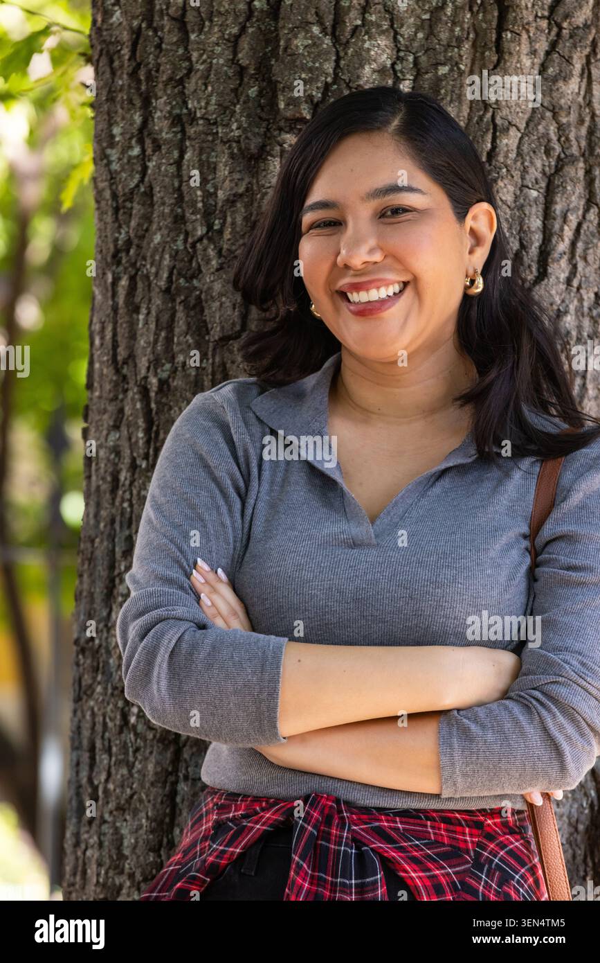 Donna adulta che si appoggia e sorride contro l'albero nel parco, braccia incrociate, con piane in maglia grigia legate Foto Stock