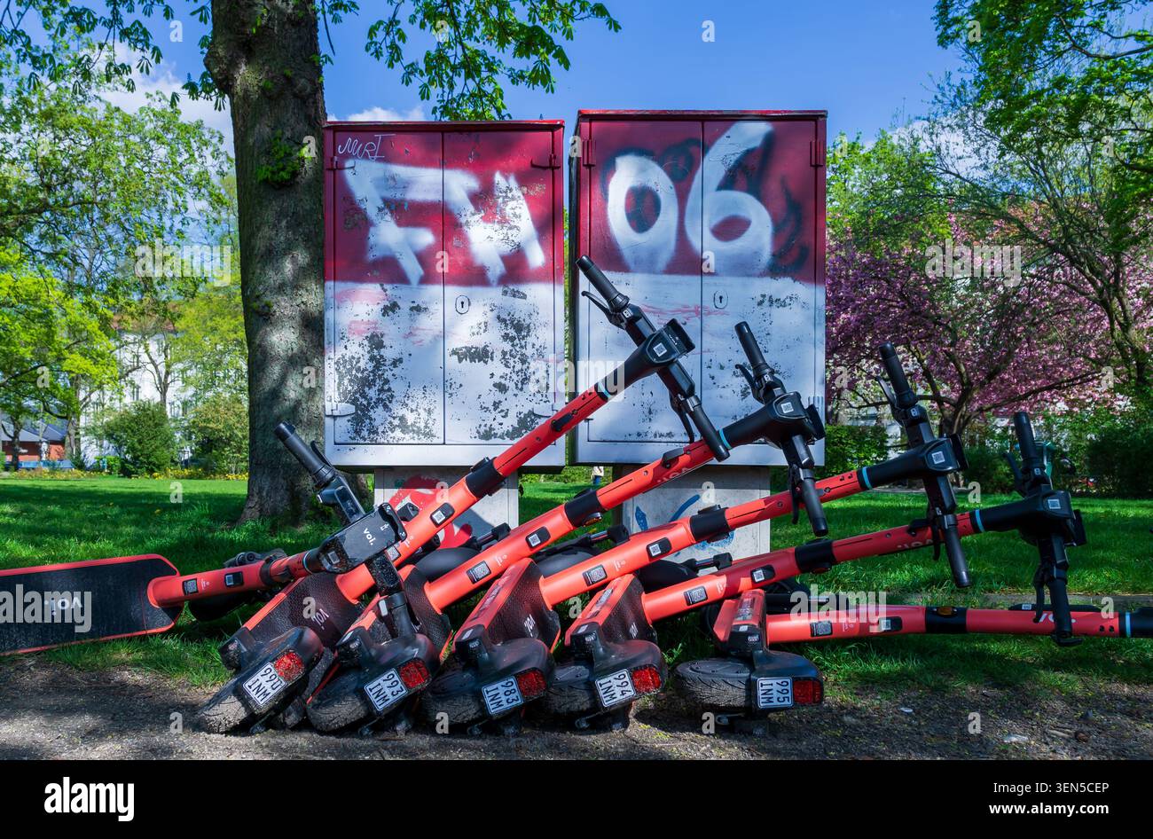 Diversi scooter elettrici condivisi sono impilati a terra di fronte a scatole per utenze coperte da graffiti in un parco pubblico, uno scooter elettrico, uno scooter elettrico rosso. Foto Stock