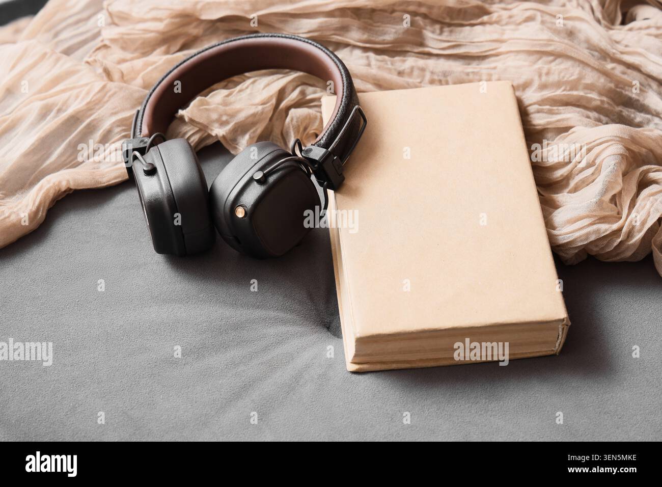 Cuffie wireless e libro bianco su tessuto grigio con sciarpa beige, apprendimento audio e relax Foto Stock