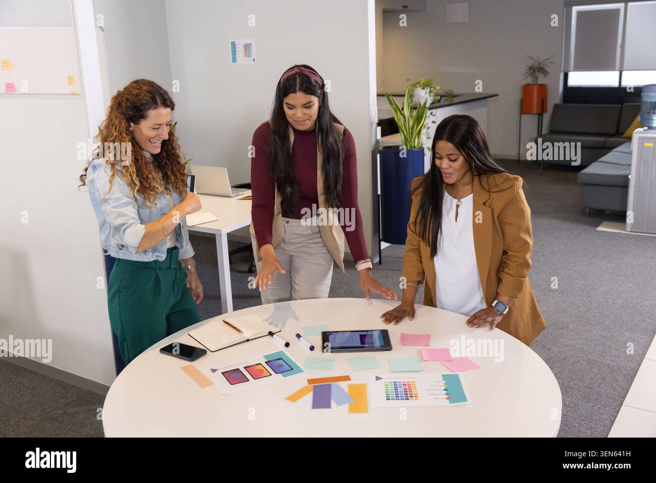 Colleghi di sesso femminile che esaminano il marchio alla tavola rotonda bianca in ufficio, utilizzando tablet e campioni a colori Foto Stock