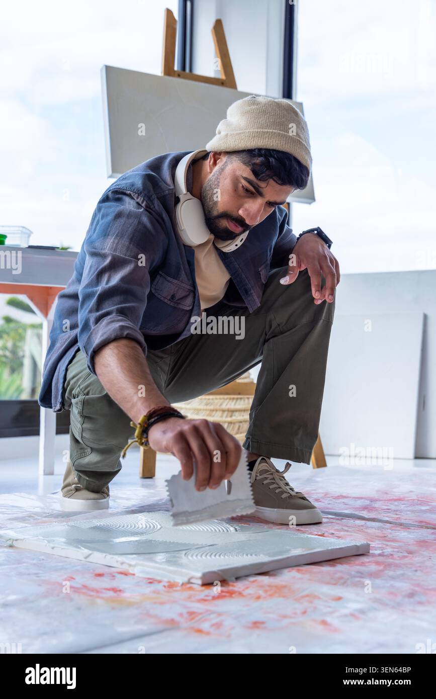 Uomo che indossa un berretto accovacciato su un panno in goccia verniciato in studio utilizzando una cazzuola intagliata sul pannello di texture Foto Stock