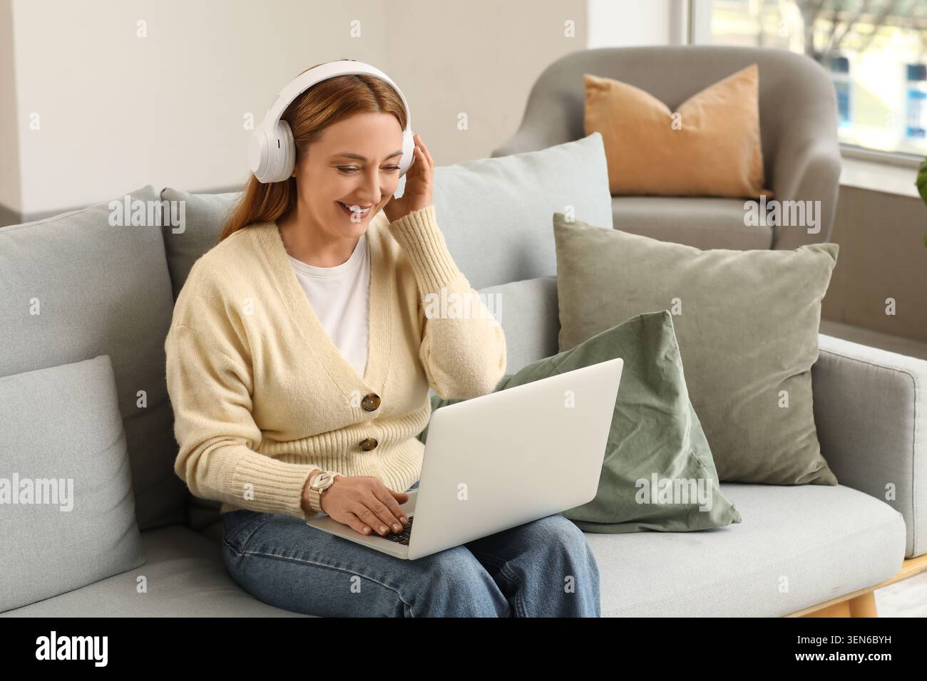 Donna sorridente che indossa le cuffie sul divano con un laptop, un lavoro remoto e uno stile di vita di apprendimento online, la postura rilassata della donna e l'espressione concentrata c Foto Stock