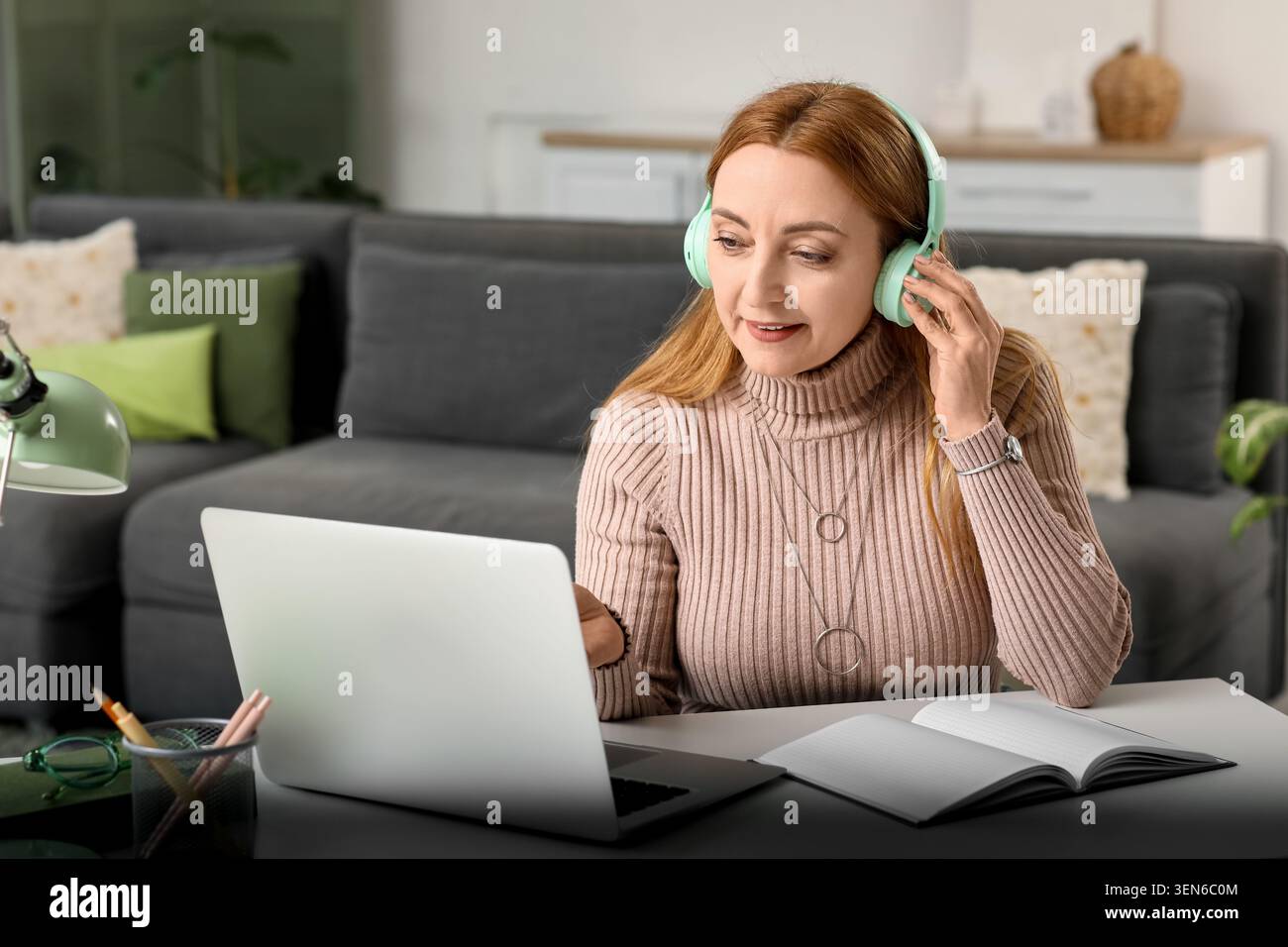 Donna matura con cuffie che lavora in remoto su un notebook dall'ufficio domestico, dall'apprendimento online e dal telelavoro Foto Stock