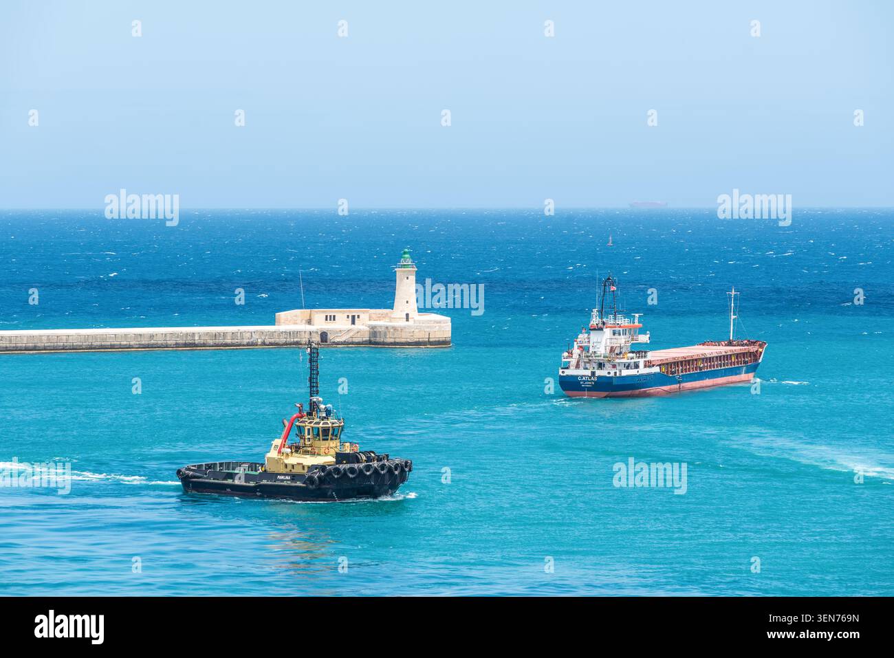 La Valletta, Malta - 16 maggio 2025: Nave da carico e rimorchiatore in barca a vela vicino allo storico ingresso del porto e al faro di la Valletta, Malta, circondato da bricco Foto Stock