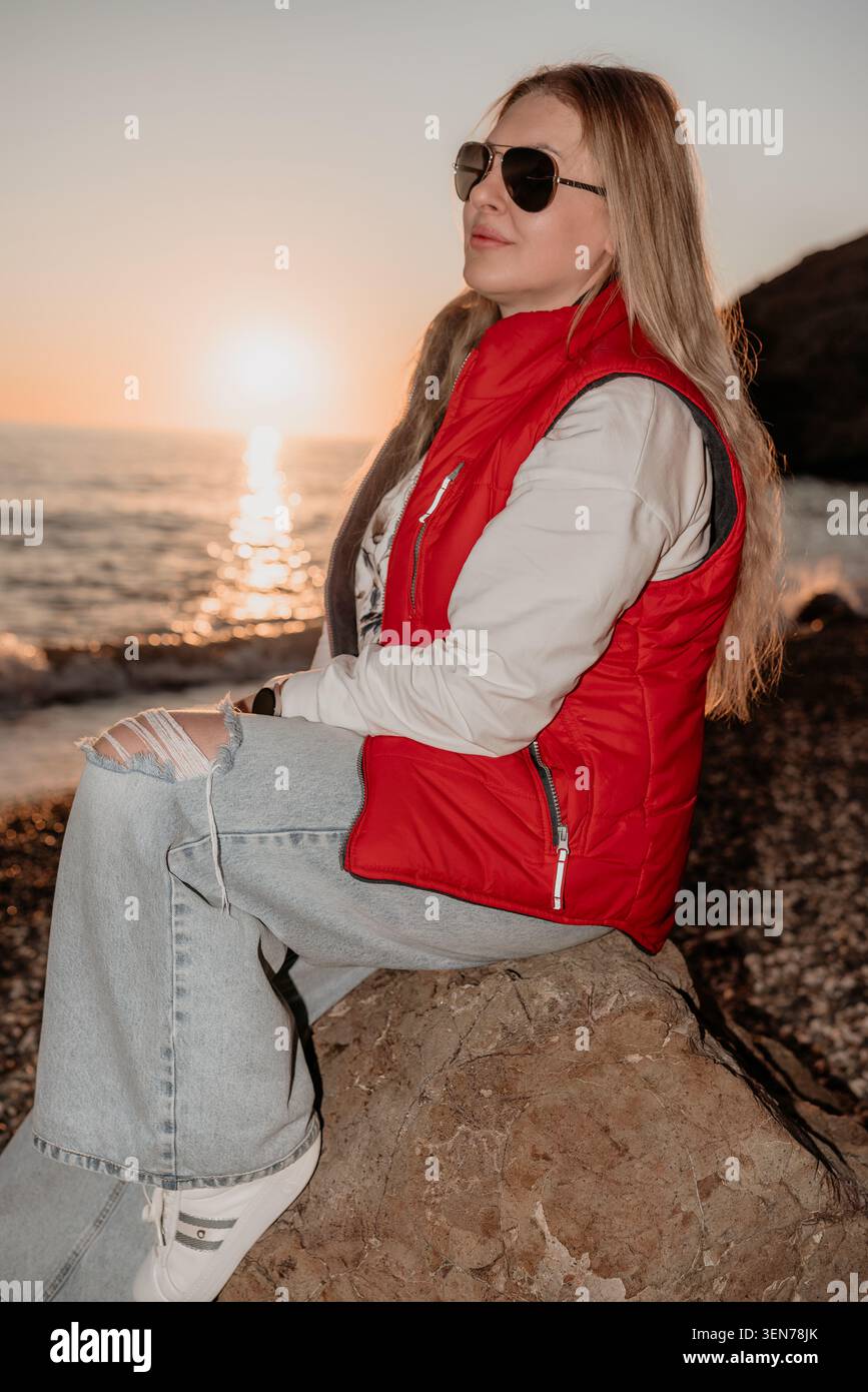 Donna al tramonto sulla spiaggia seduta su una roccia sulla riva con un gilet rosso e occhiali da sole godendosi il bagliore della sera Foto Stock